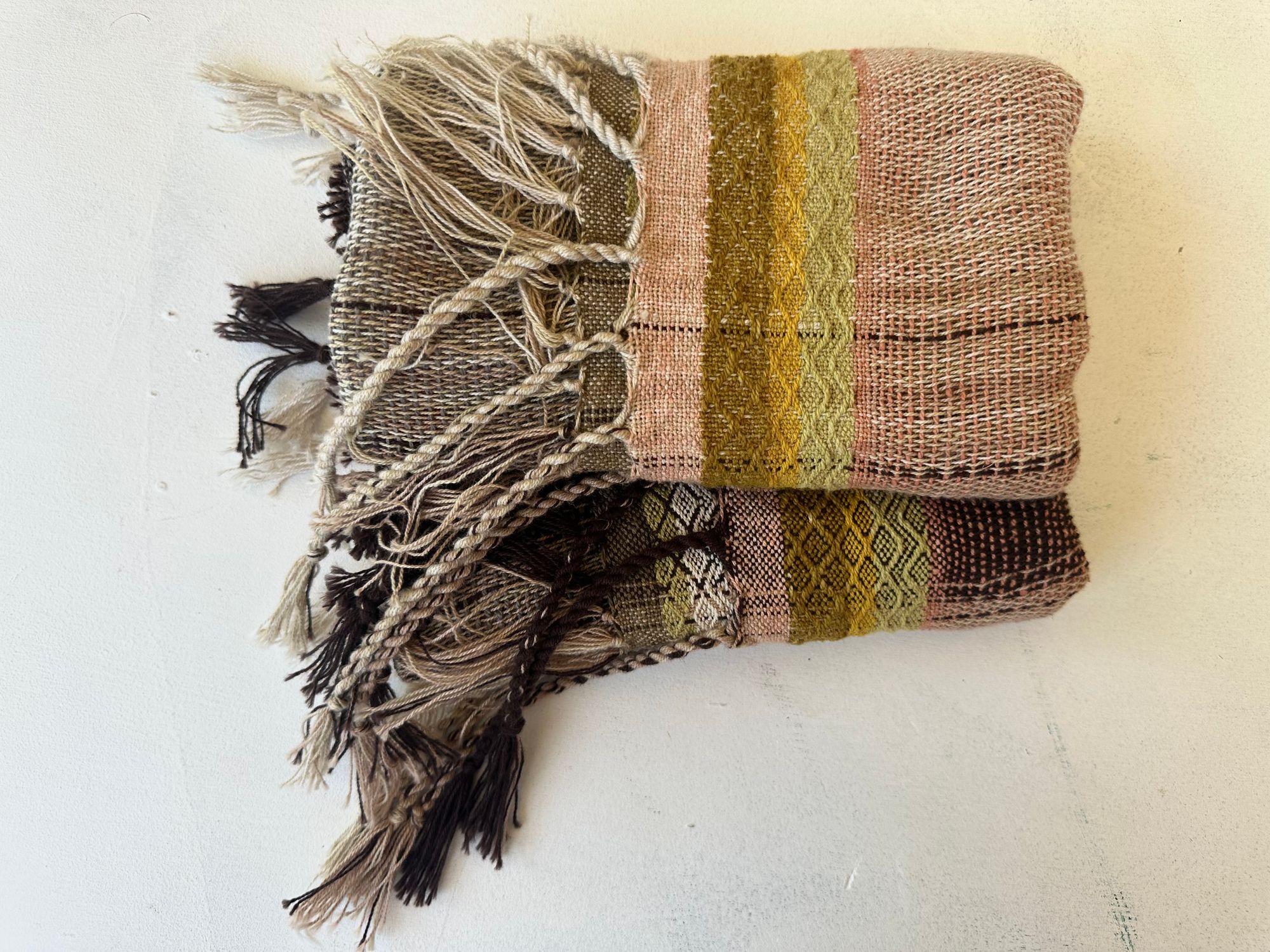 Hand woven scarf in brown, yellows and soft pinks is folded on a white floor