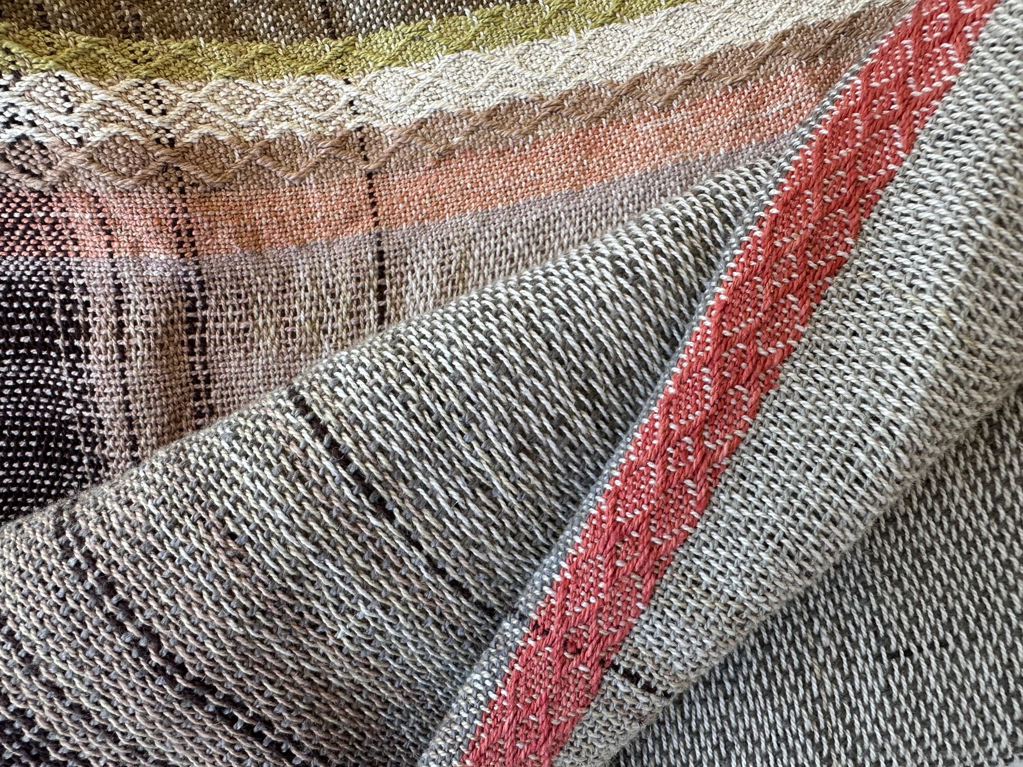 A detail of Hand woven scarf in brown, yellows and soft pinks is folded on a white floor