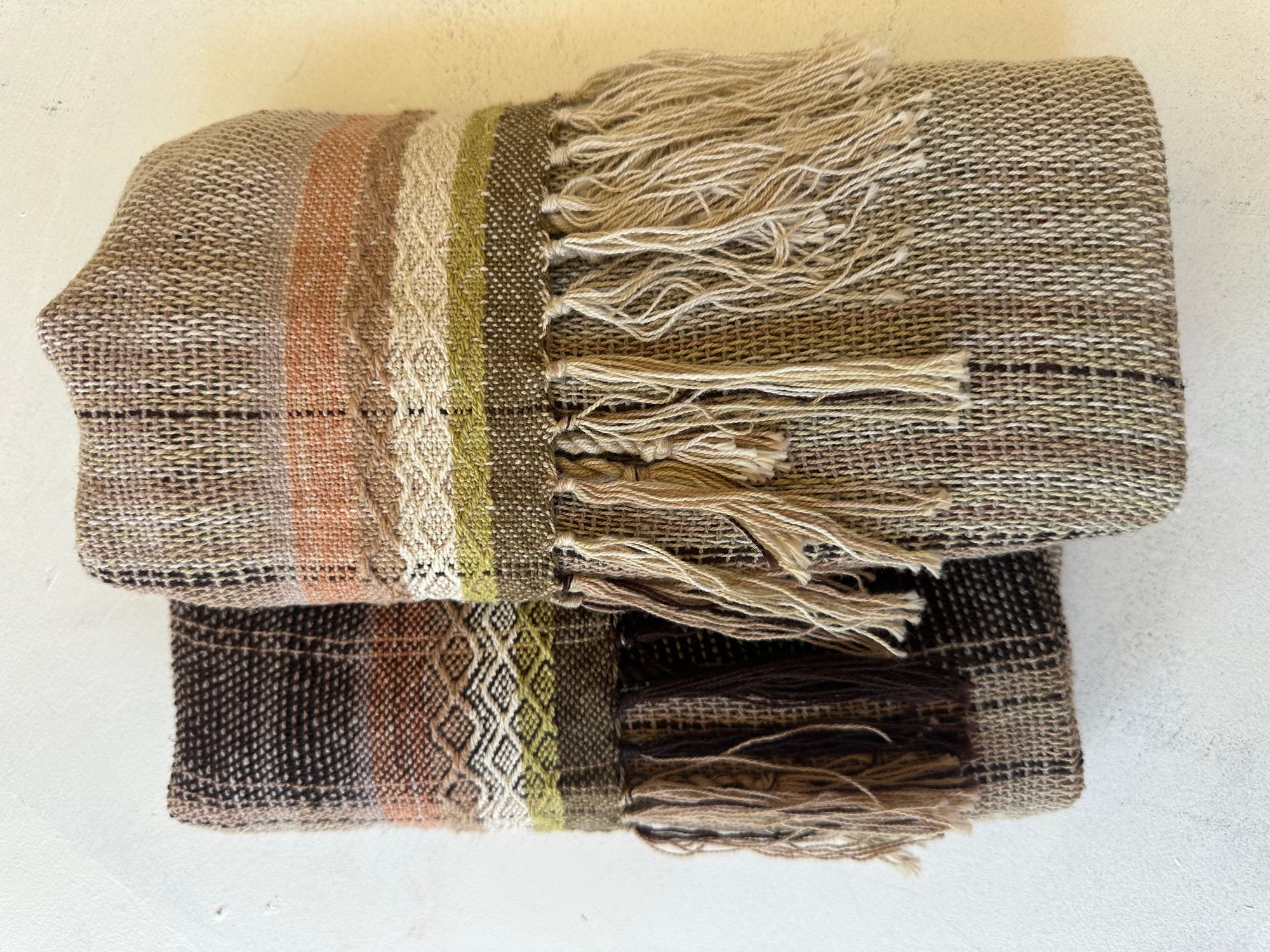 Hand woven scarf in brown, yellows and soft pinks is folded on a white floor