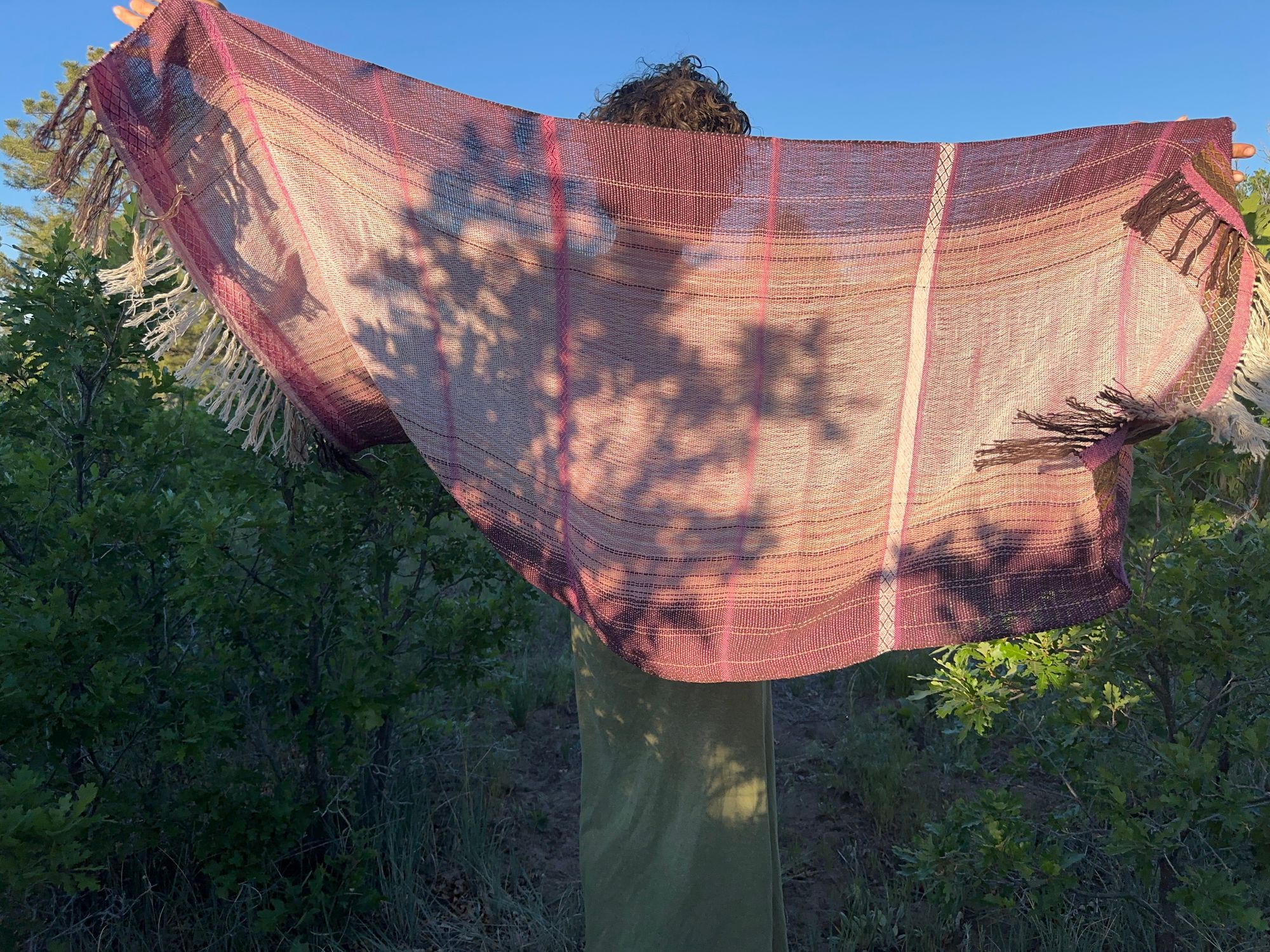 A woman in a brown shirt and green skirt wears a handwoven Naturally dyed pink shawl in the forest