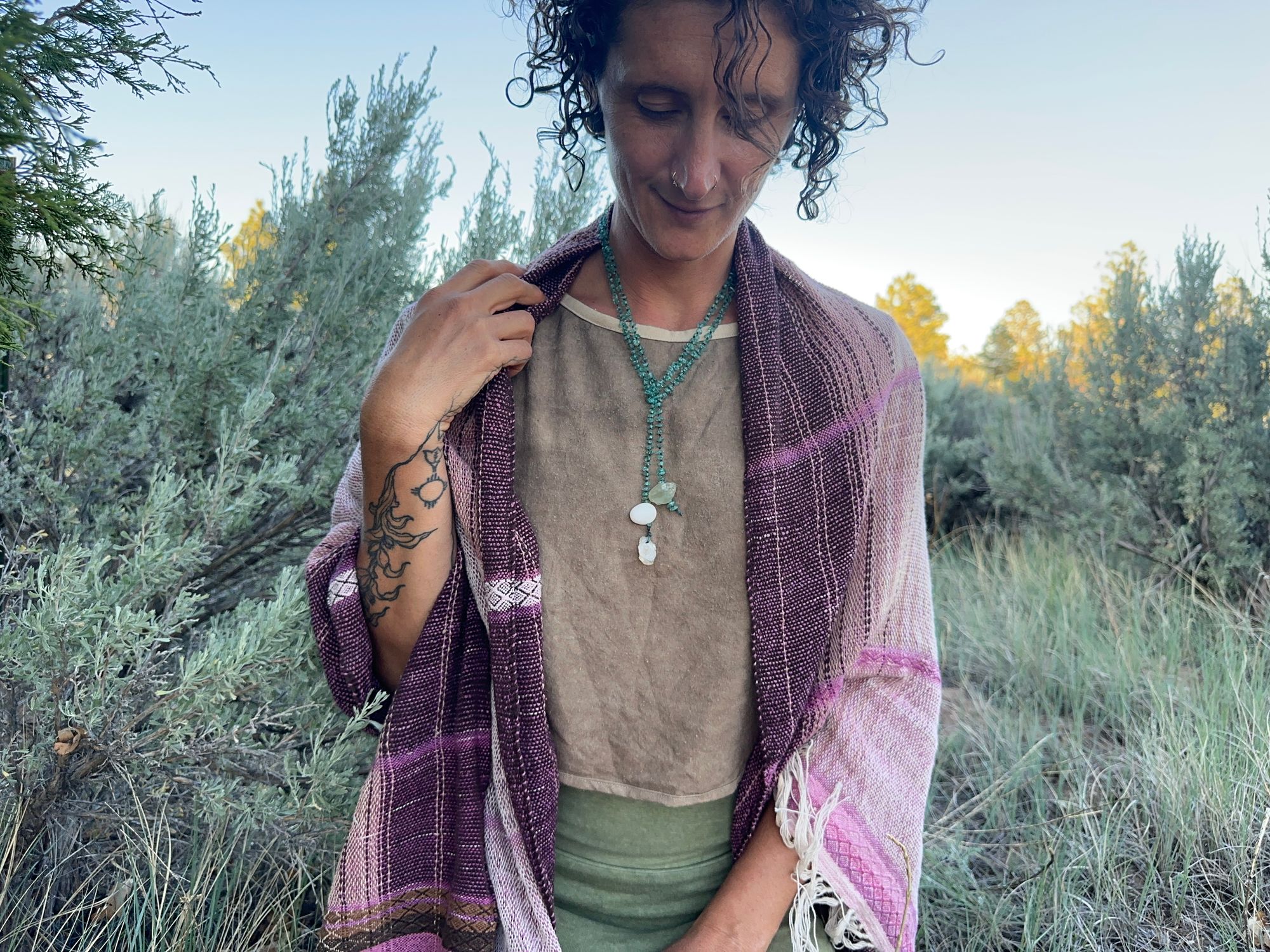 A woman in a brown shirt and green skirt wears a handwoven Naturally dyed pink shawl in the forest