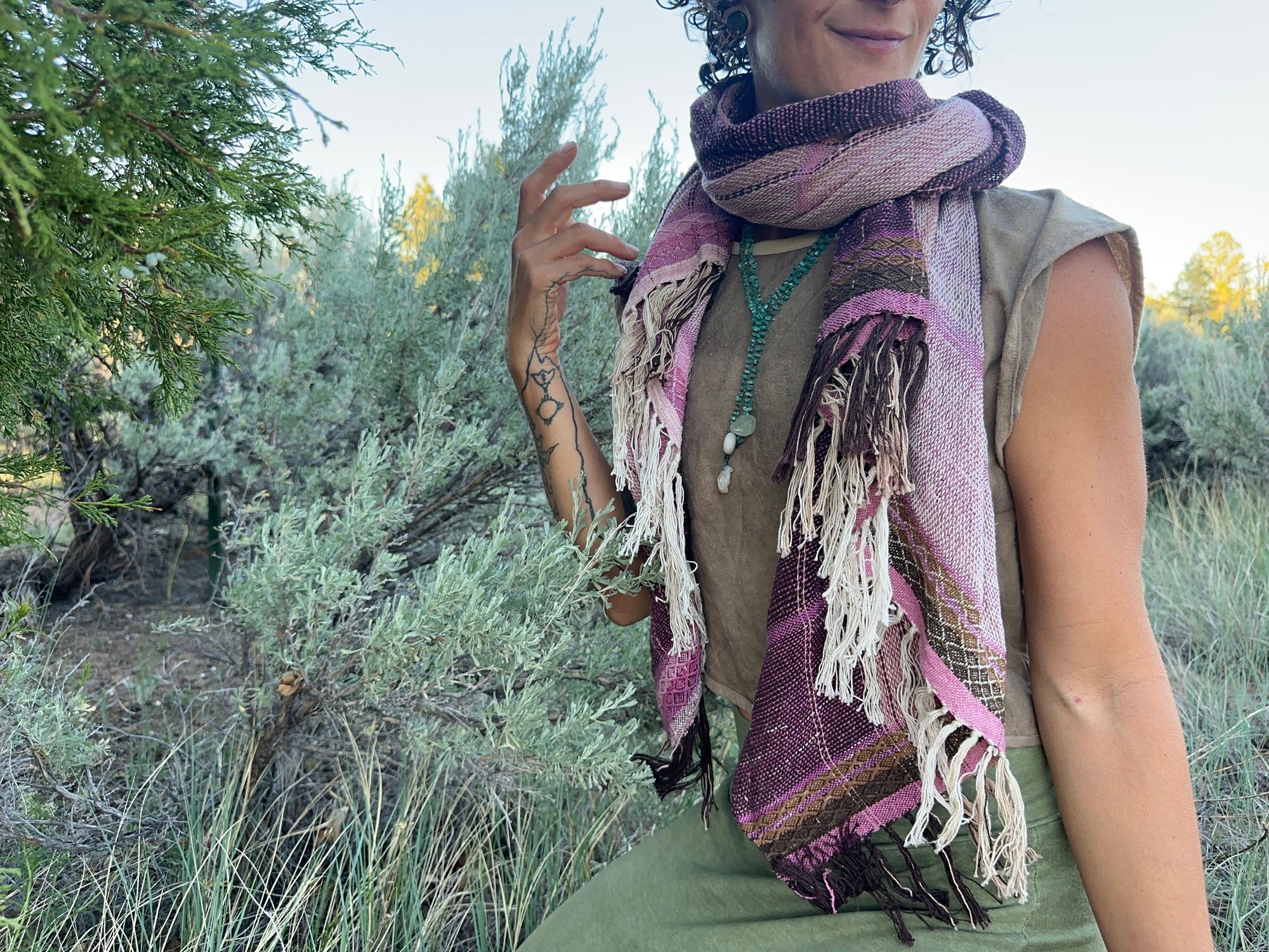 A woman in a brown shirt and green skirt wears a handwoven Naturally dyed pink shawl in the forest