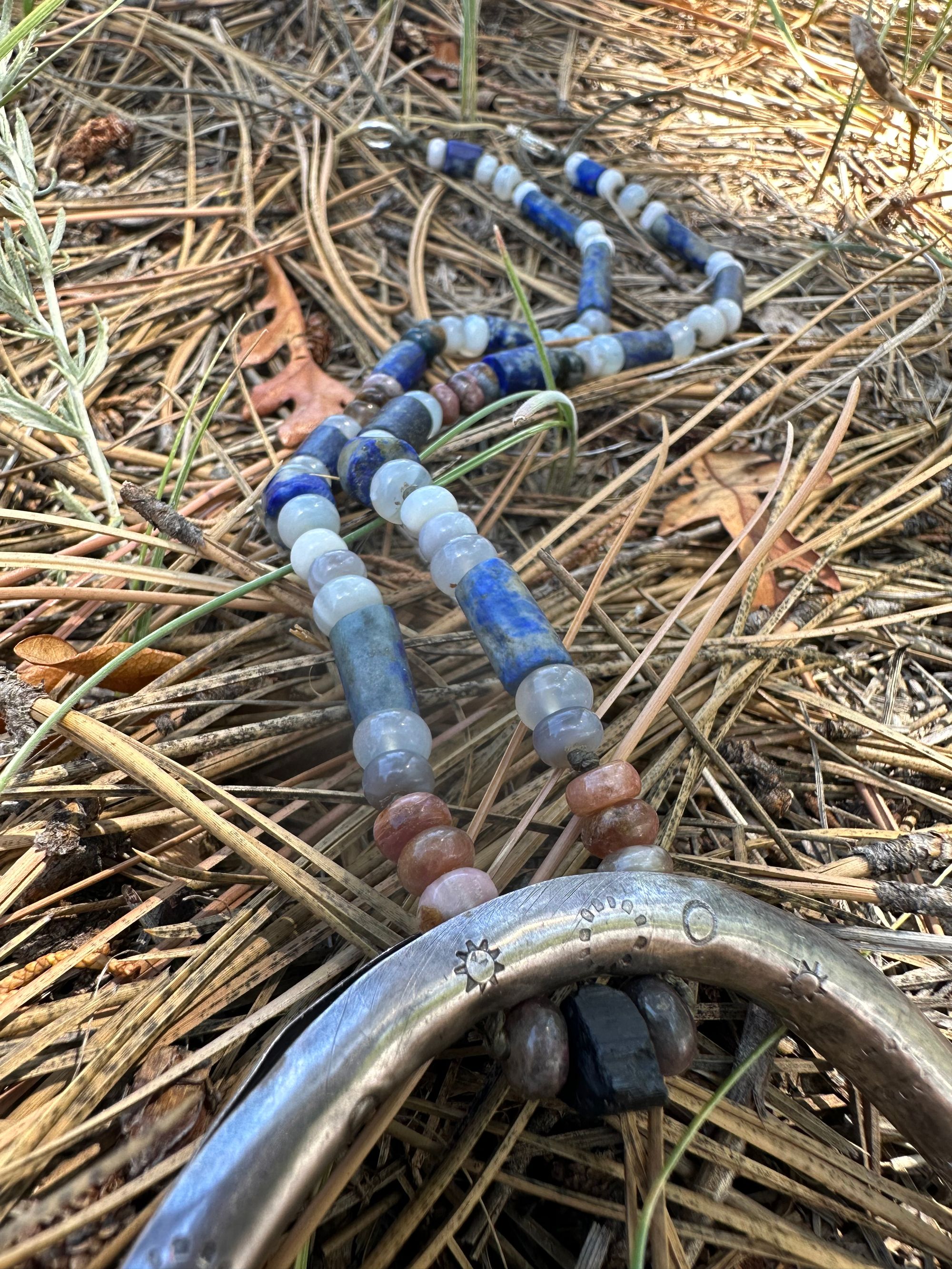 A necklace made of semi precious stones and a solid silver moon lays on Pineneedles