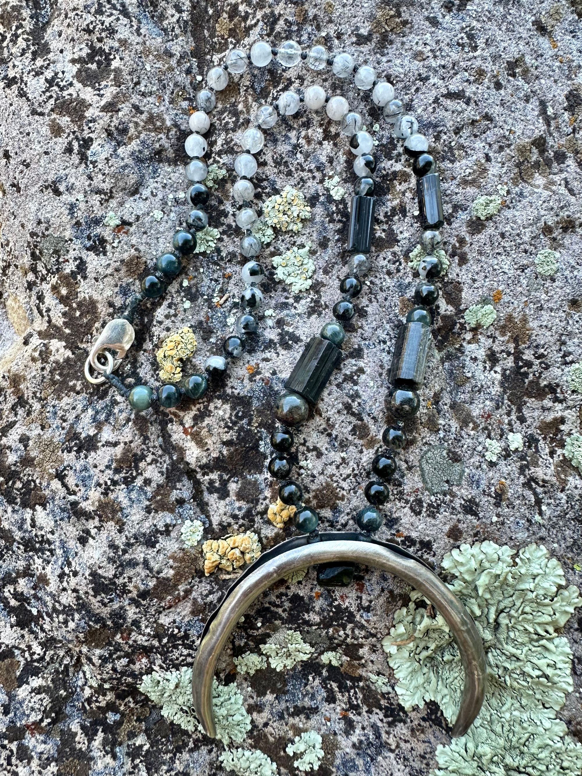 A necklace made of semi precious stones and a solid silver moon lays on a lichen covered rock