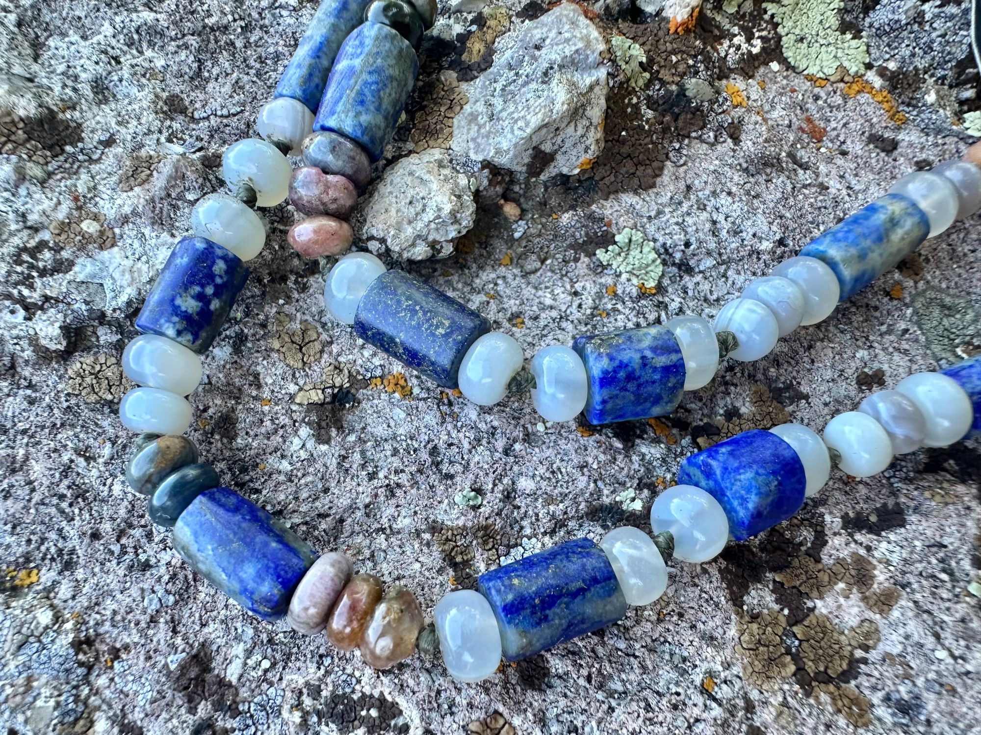 A necklace made of semi precious stones and a solid silver moon lays on a lichen covered rock