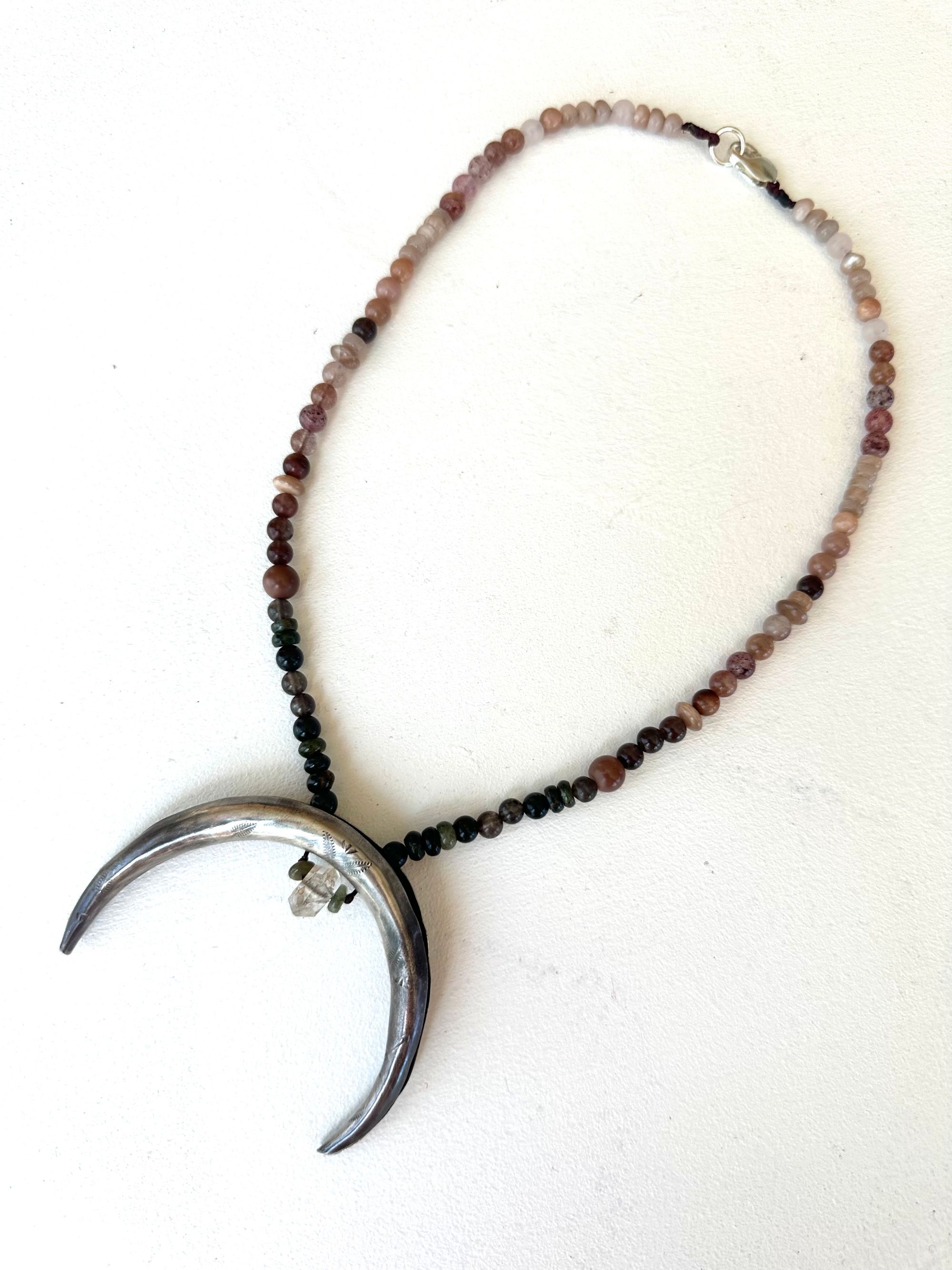 A necklace made of a solid silver moon and semi precious stone beads rests on a white floor