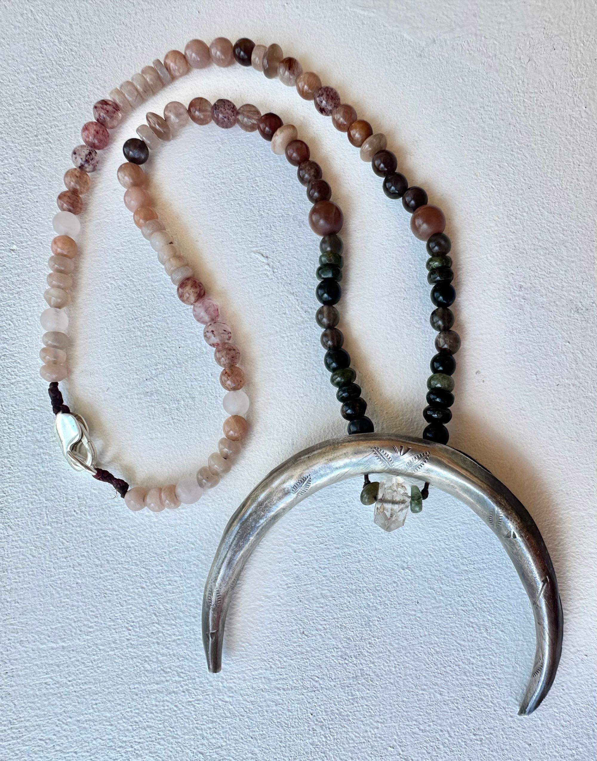 A necklace made of a solid silver moon and semi precious stone beads rests on a white floor