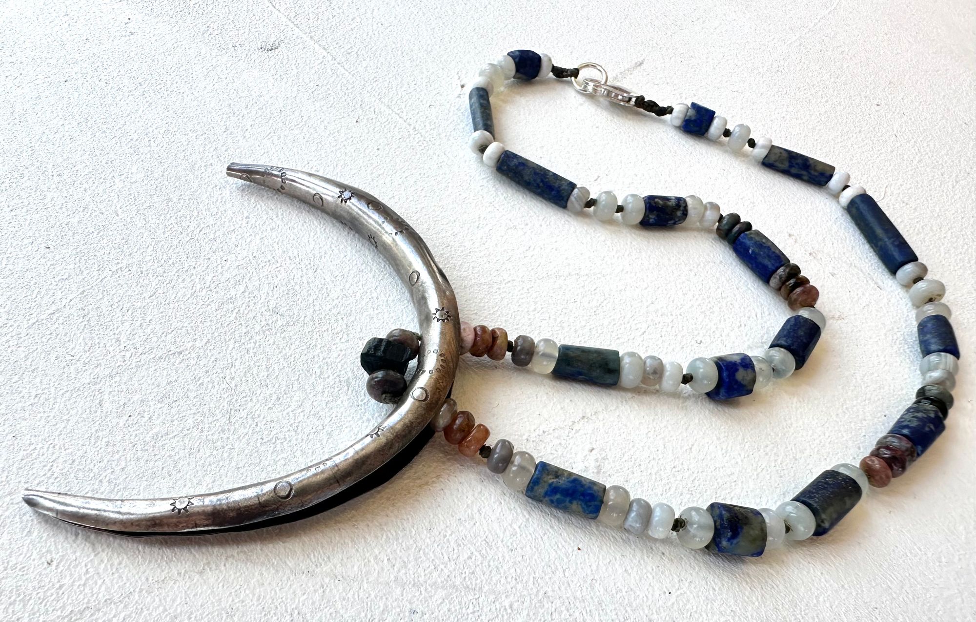 A necklace made of a solid silver moon and semi precious stone beads rests on a white floor