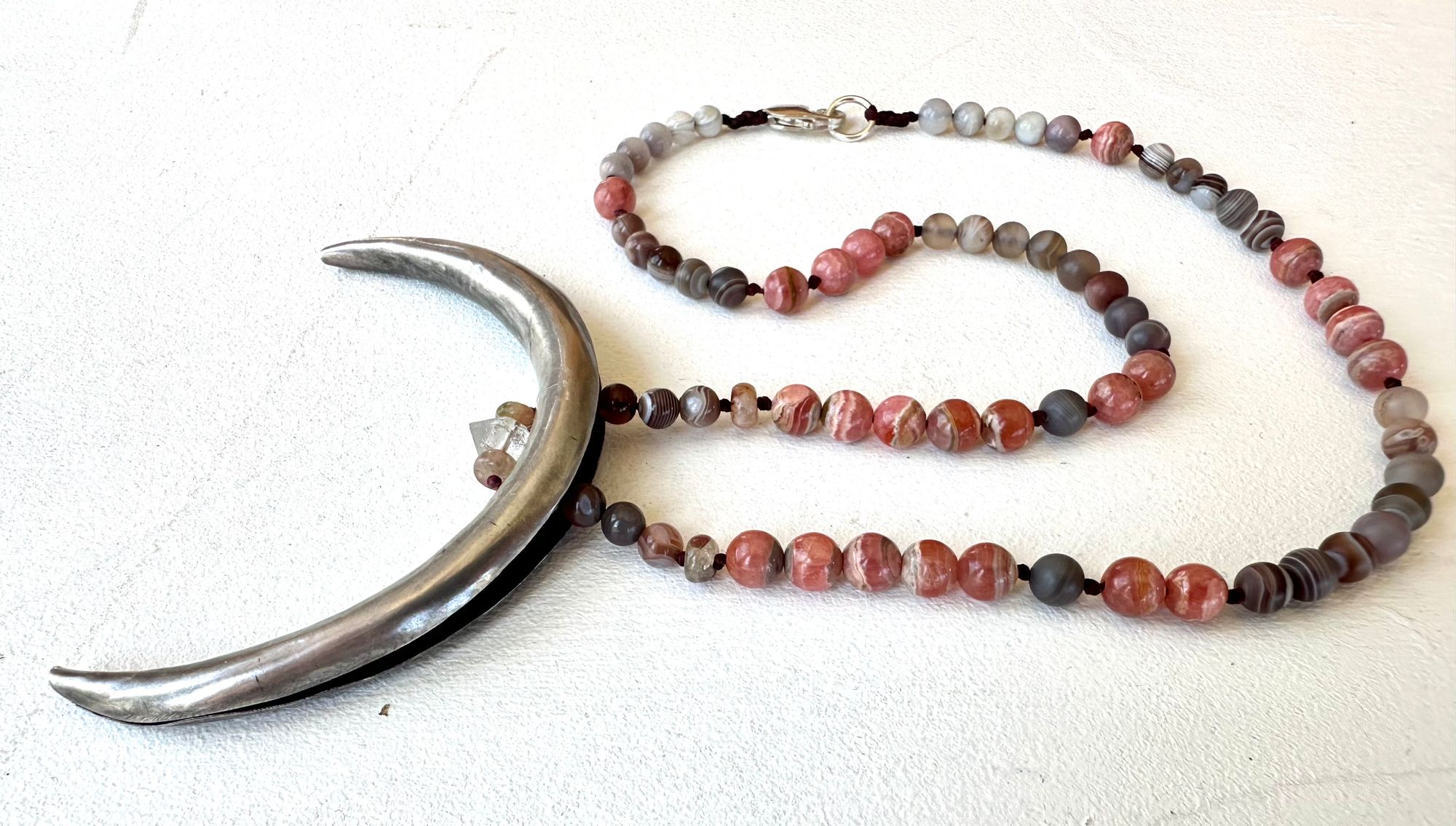 A necklace made of a solid silver moon and semi precious stone beads rests on a white floor