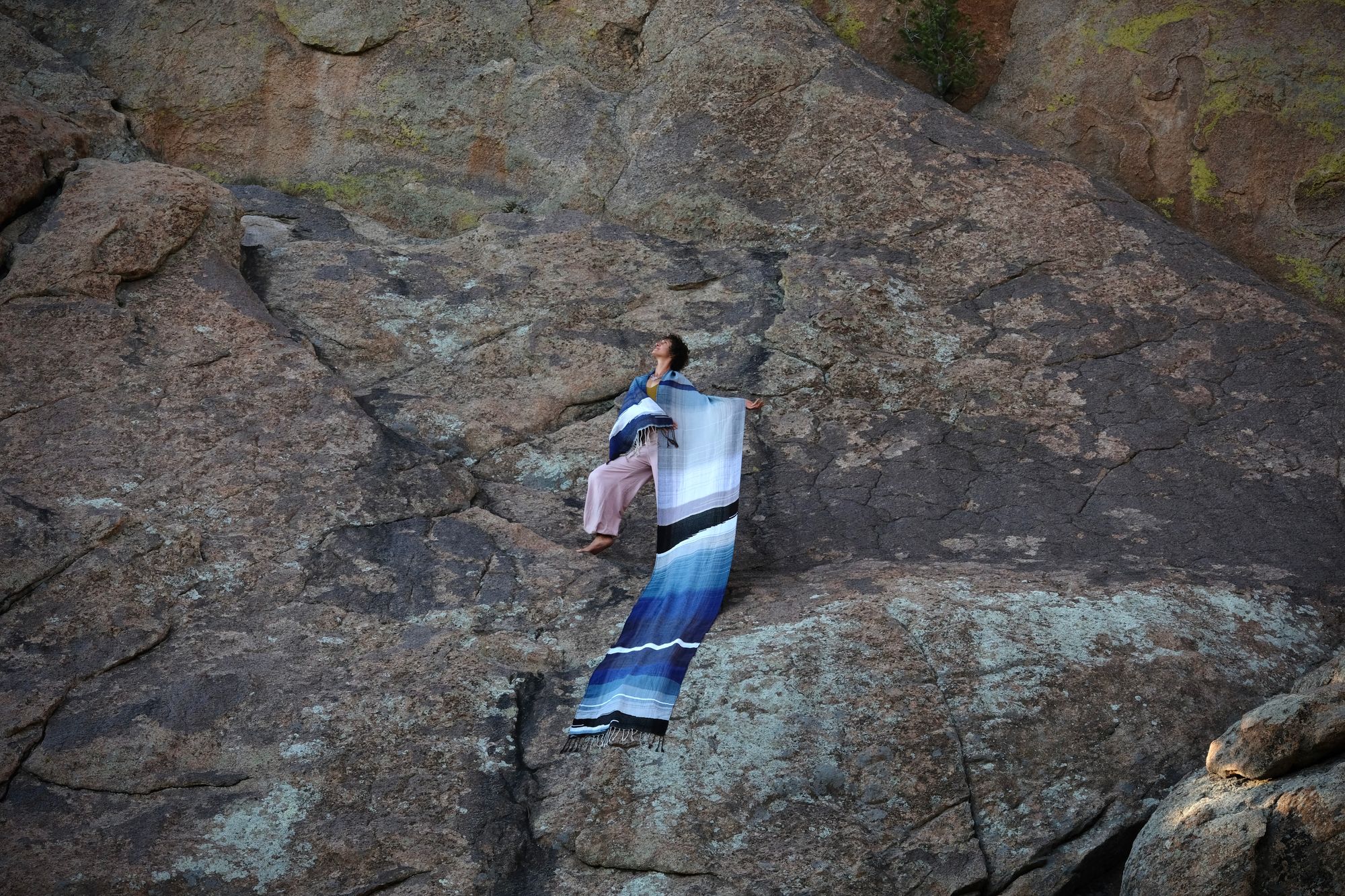 A piece of handwoven, diamond patterned, with wave-like designs fabric in shades of white, to blue, to black is held by a woman standing on a deep red-brown granite, lichen covered dome