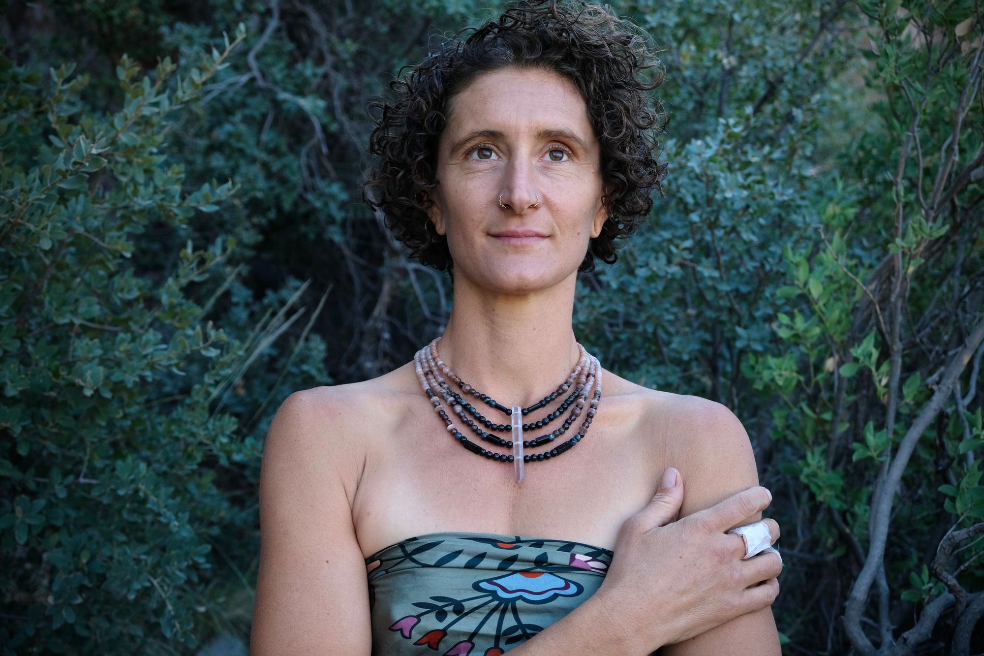 A woman wears a necklace of semi precious stones in shades of black fading to dusty Rose quartz, she wears a floral pattern handkerchief against a backdrop of dark forest