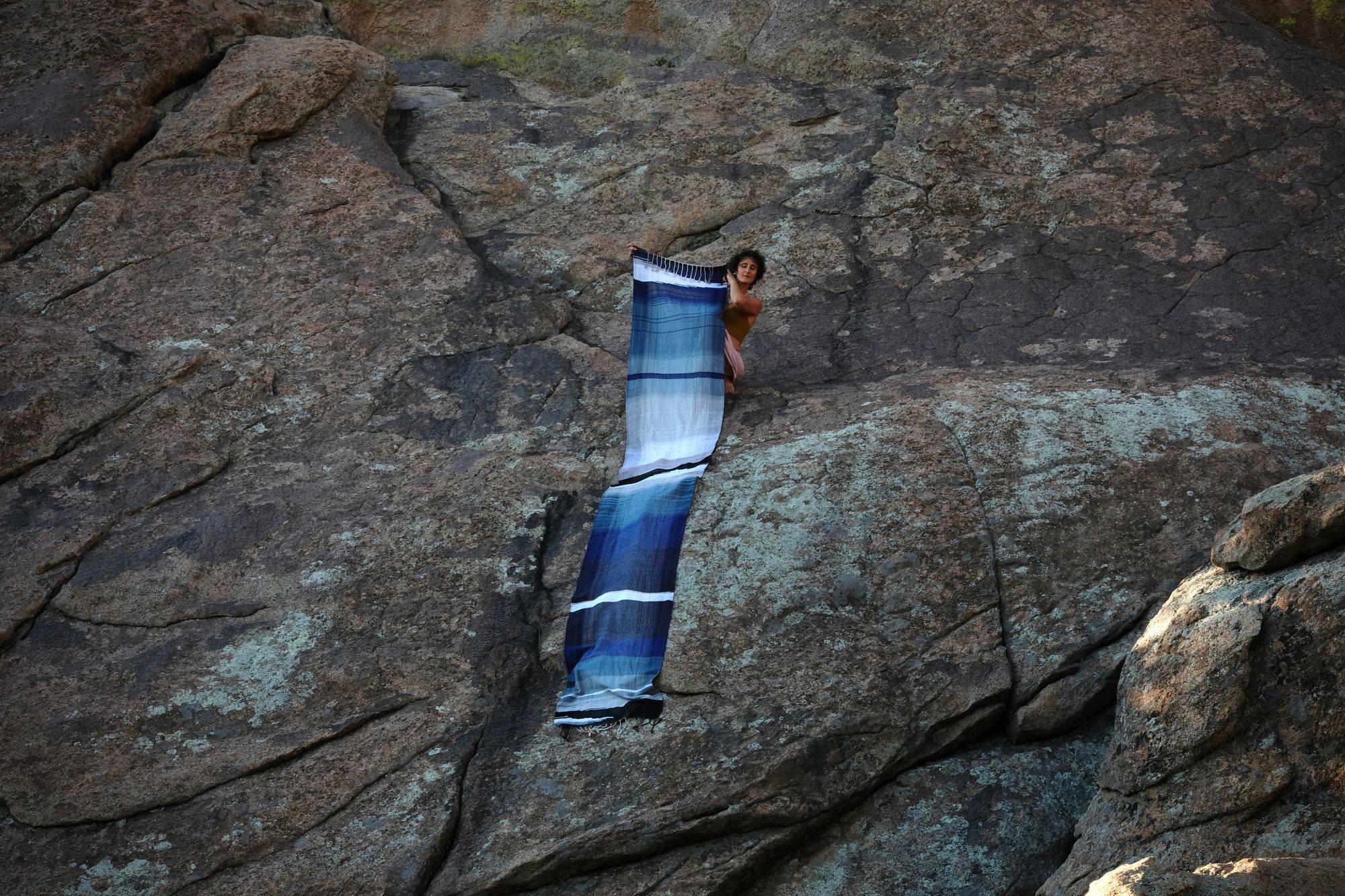 A piece of handwoven, diamond patterned, with wave-like designs fabric in shades of white, to blue, to black is held by a woman standing on a deep red-brown granite, lichen covered dome