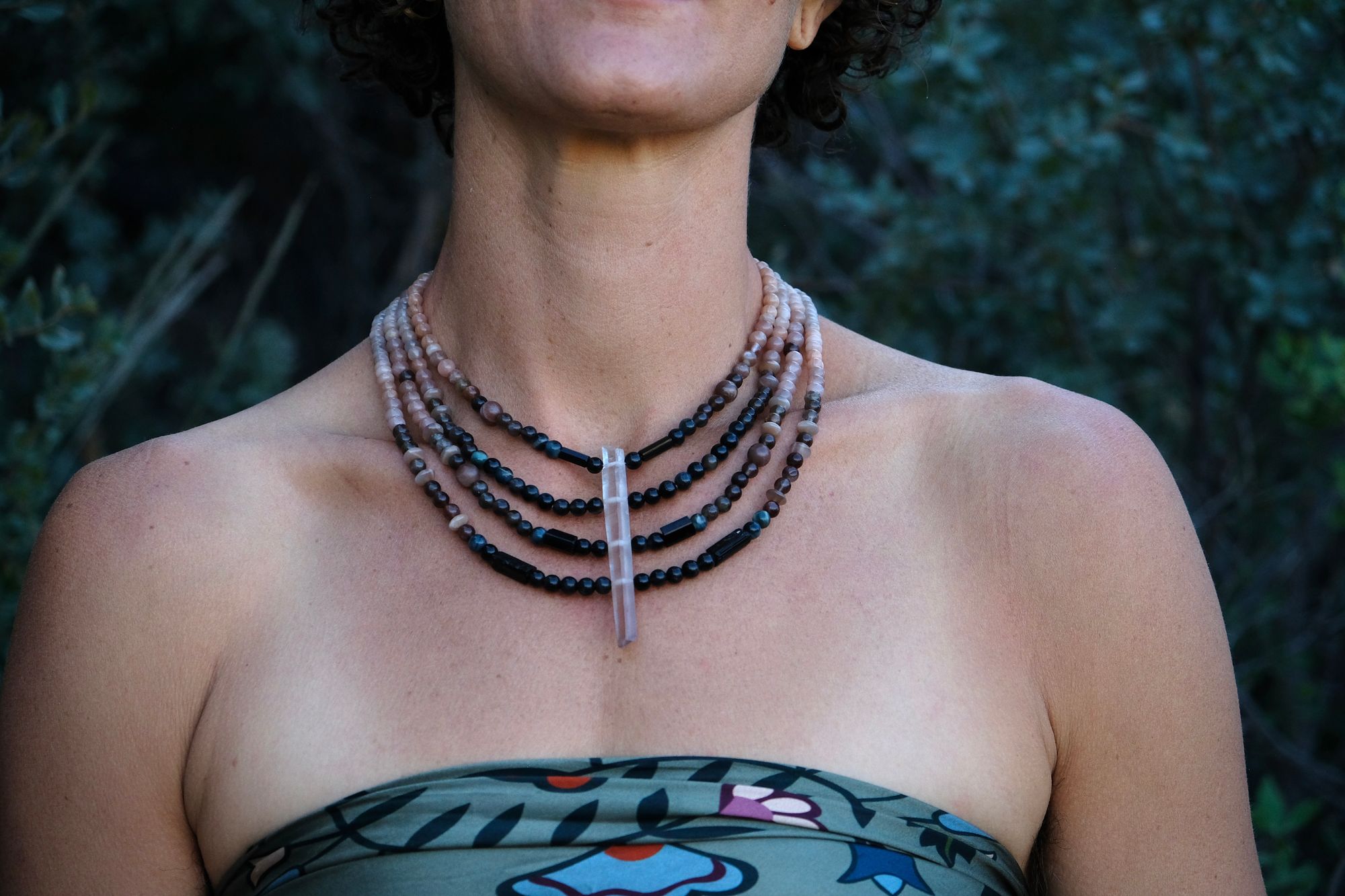 A woman wears a necklace of semi precious stones in shades of black fading to dusty Rose quartz, she wears a floral pattern handkerchief against a backdrop of dark forest