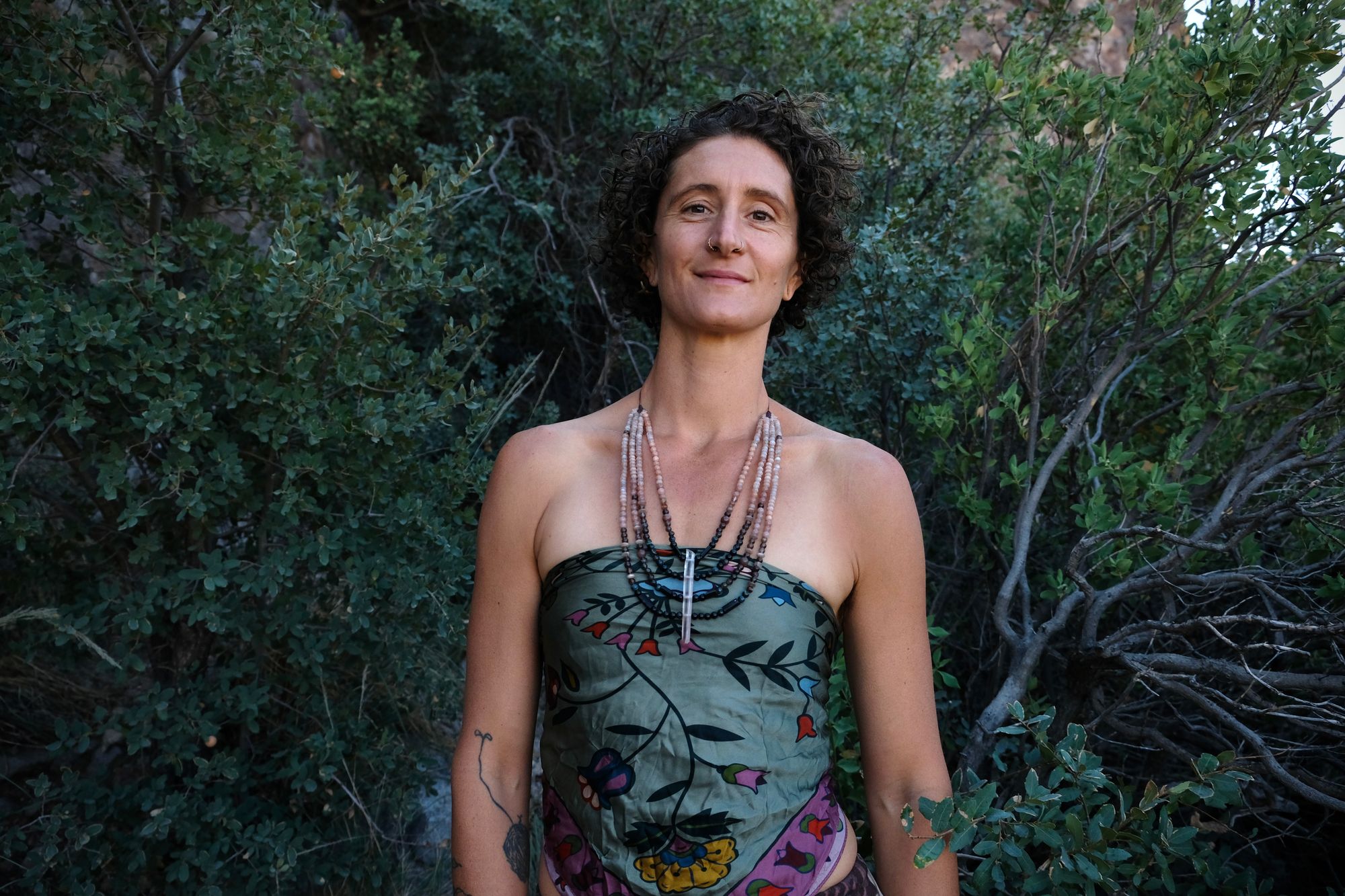 A woman wears a necklace of semi precious stones in shades of black fading to dusty Rose quartz, she wears a floral pattern handkerchief against a backdrop of dark forest