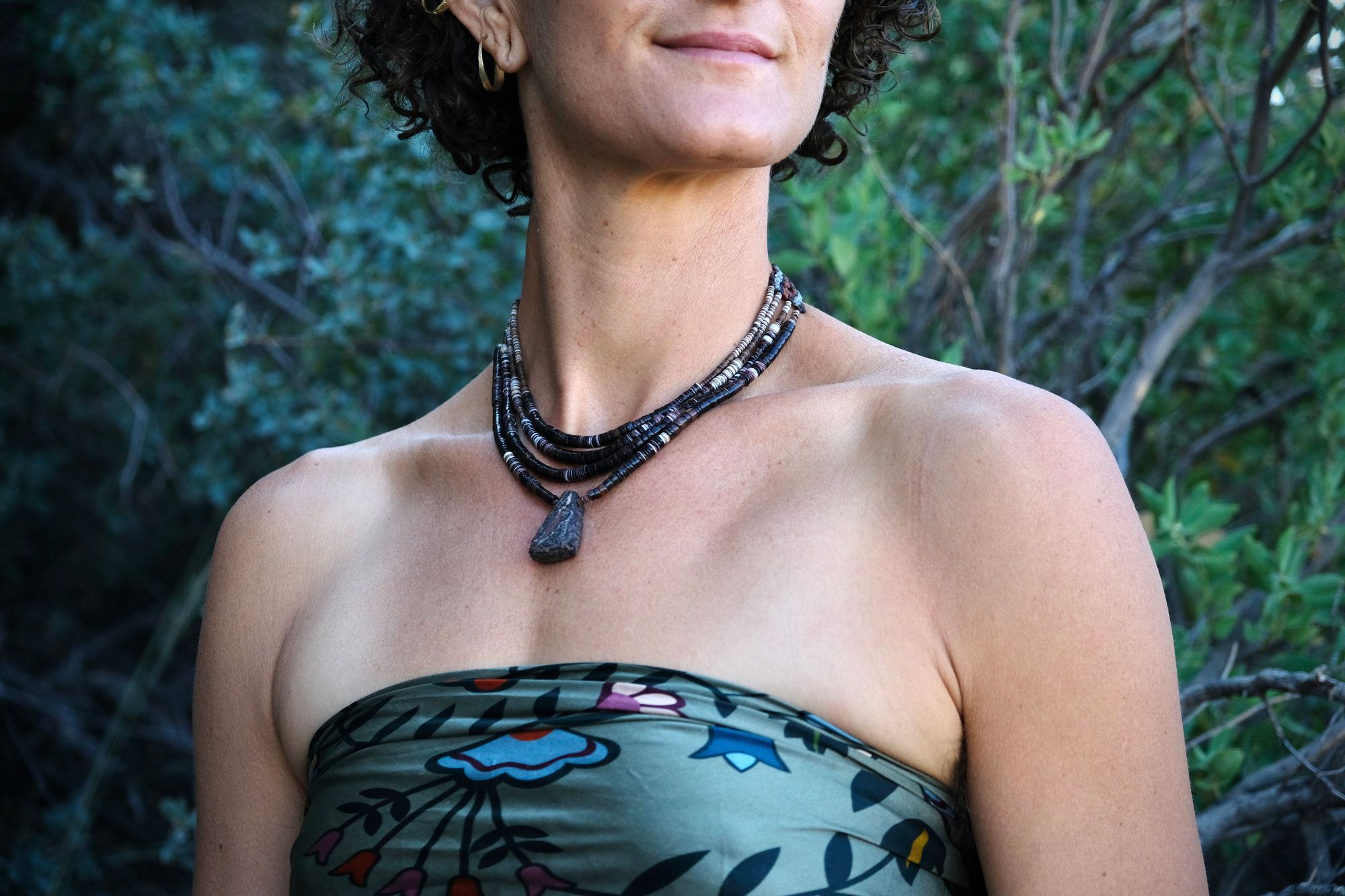 A woman wears a necklace of dark shell beads against a dark wooded background