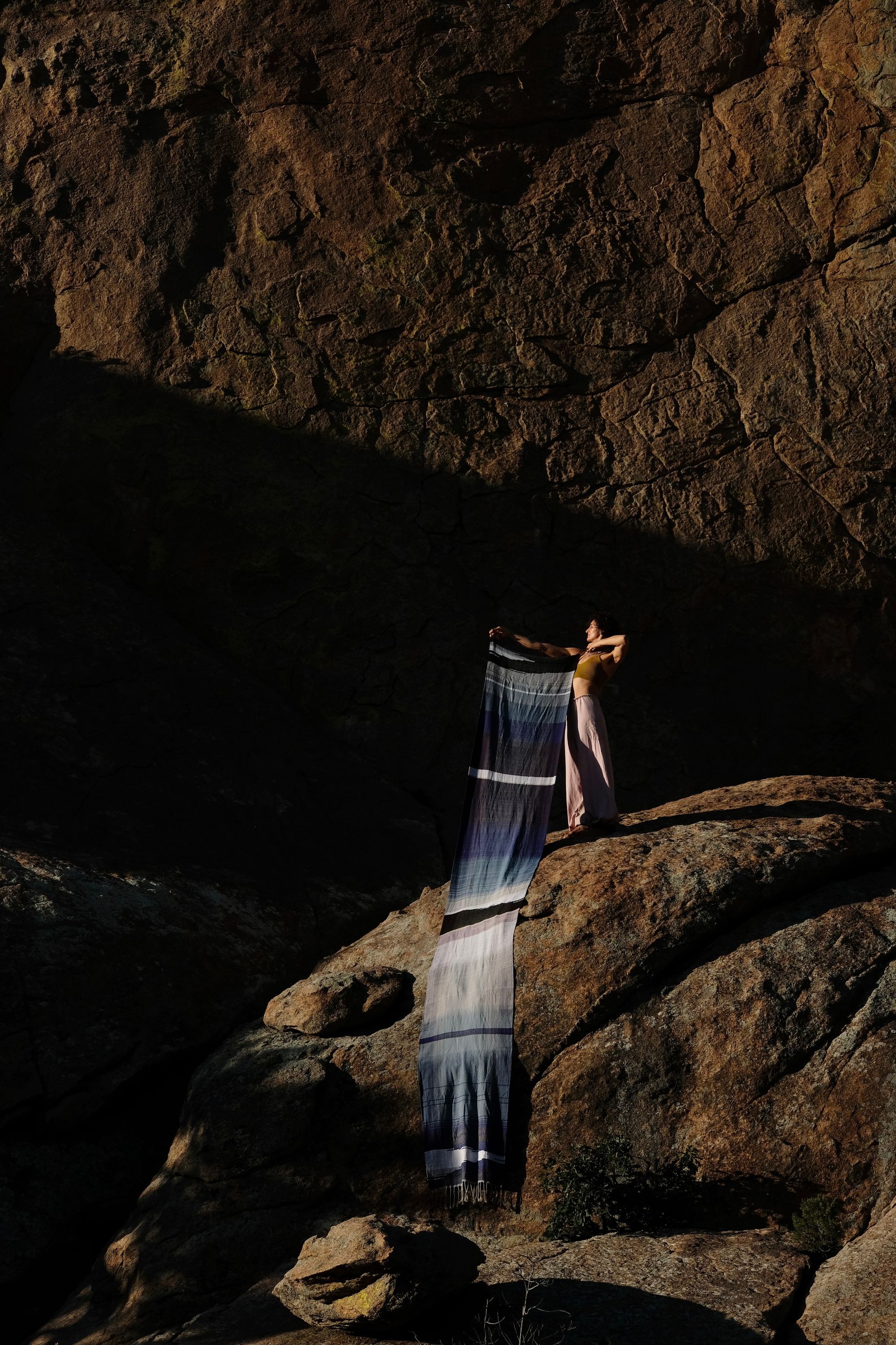 A piece of handwoven, diamond patterned, with wave-like designs fabric in shades of white, to blue, to black is held by a woman standing on a deep red-brown granite, lichen covered dome