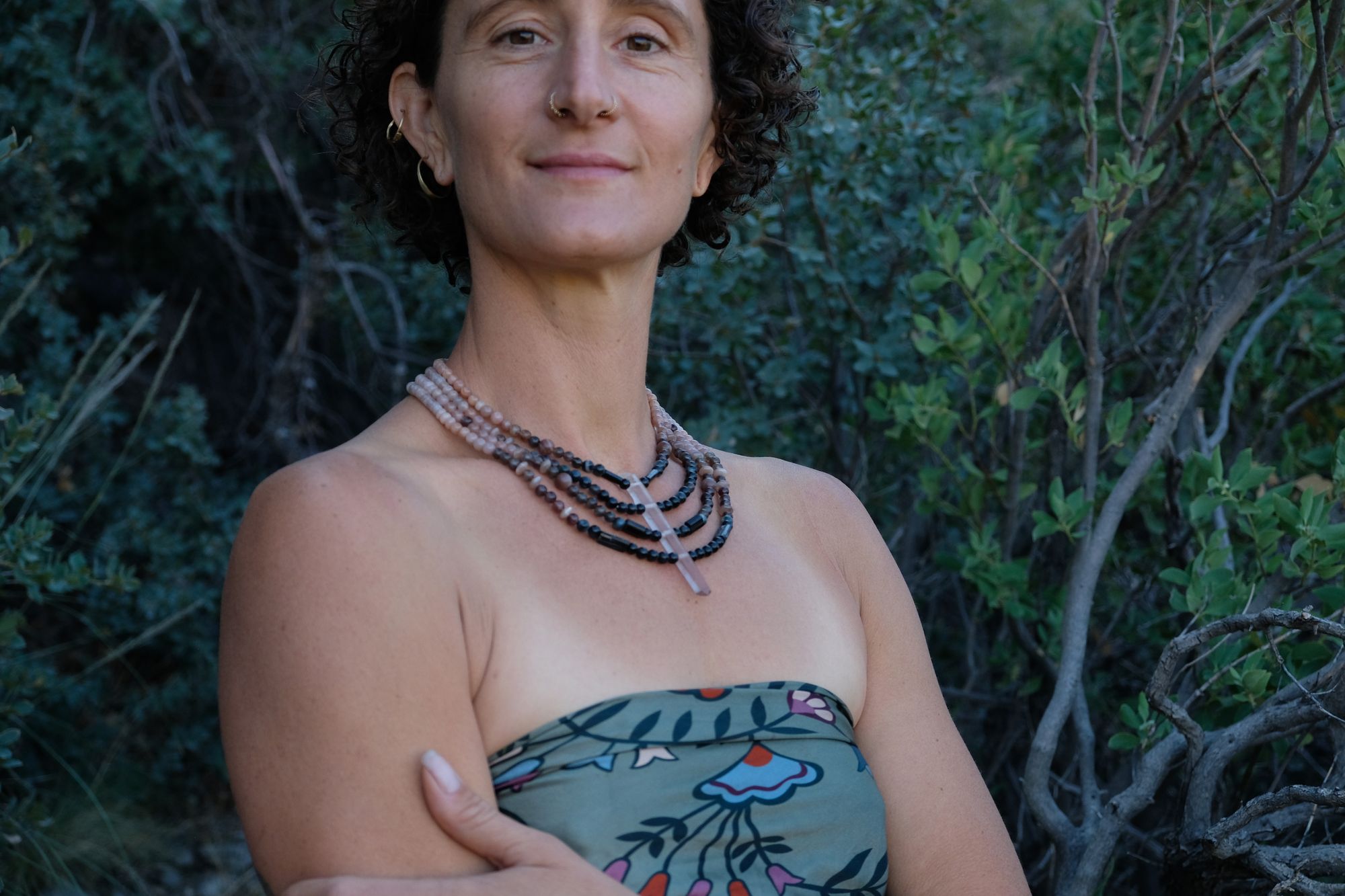 A woman wears a necklace of semi precious stones in shades of black fading to dusty Rose quartz, she wears a floral pattern handkerchief against a backdrop of dark forest