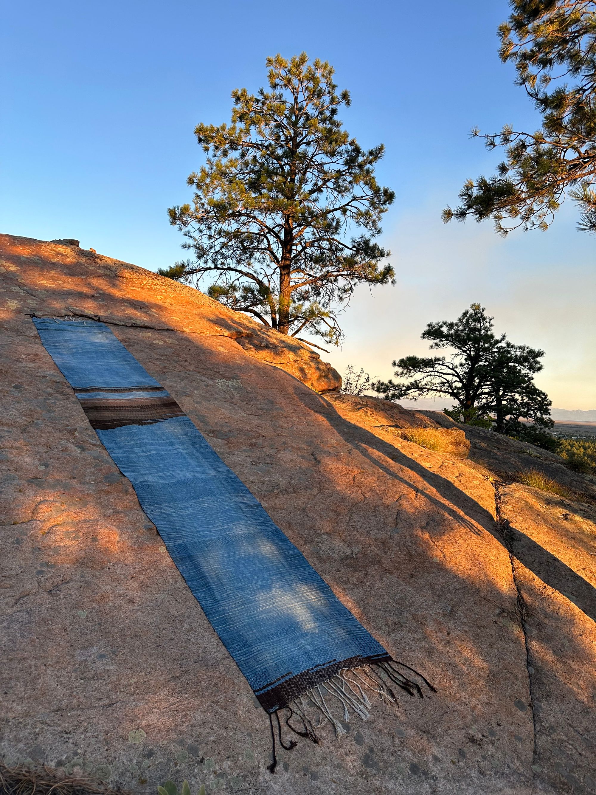 a 4.6 meter piece of handwoven fabric that is indigo blue and deep walnut dyed brown lays on a large granite stone bathed in sunset light