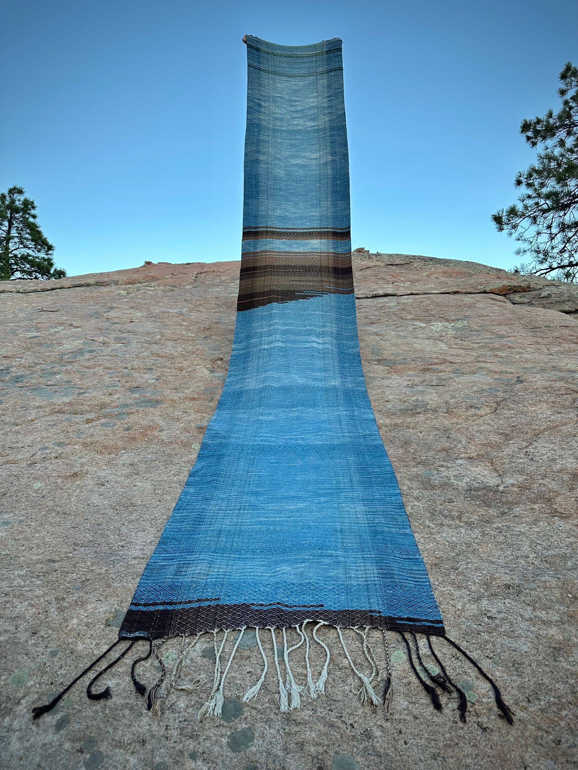 a 4.6 meter piece of handwoven fabric that is indigo blue and deep walnut dyed brown is held above a large lichen covered boulder