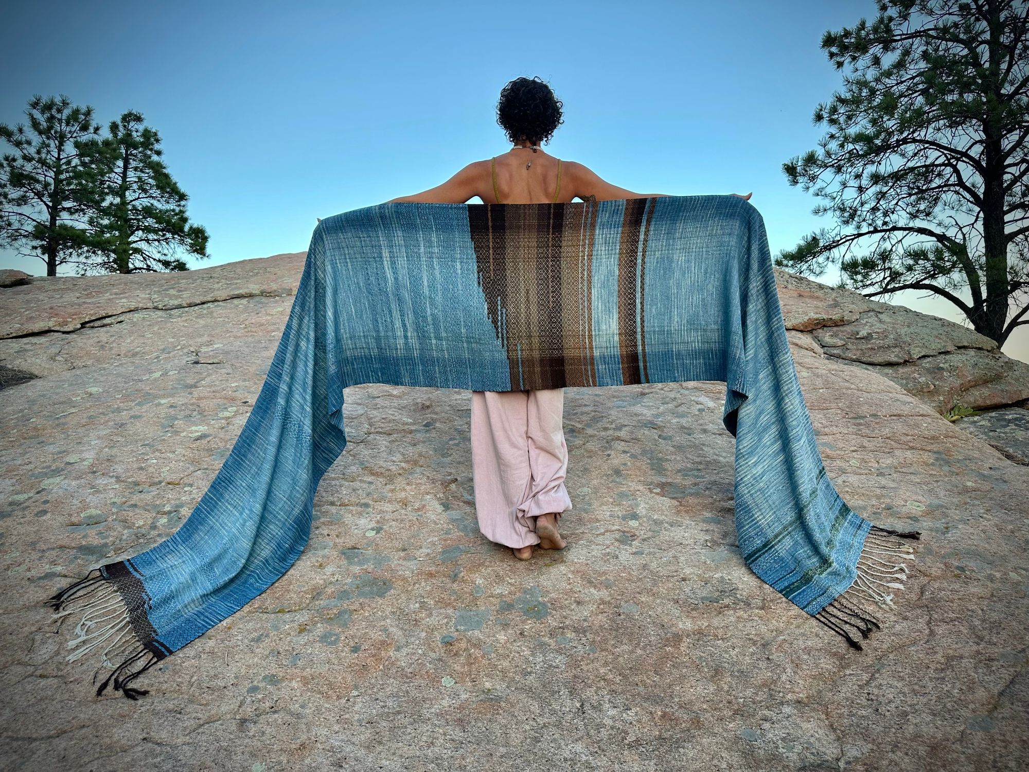 a 4.6 meter piece of handwoven fabric that is indigo blue and deep walnut dyed brown wrapped around the shoulders of a woman standing on a giant monolithic stone