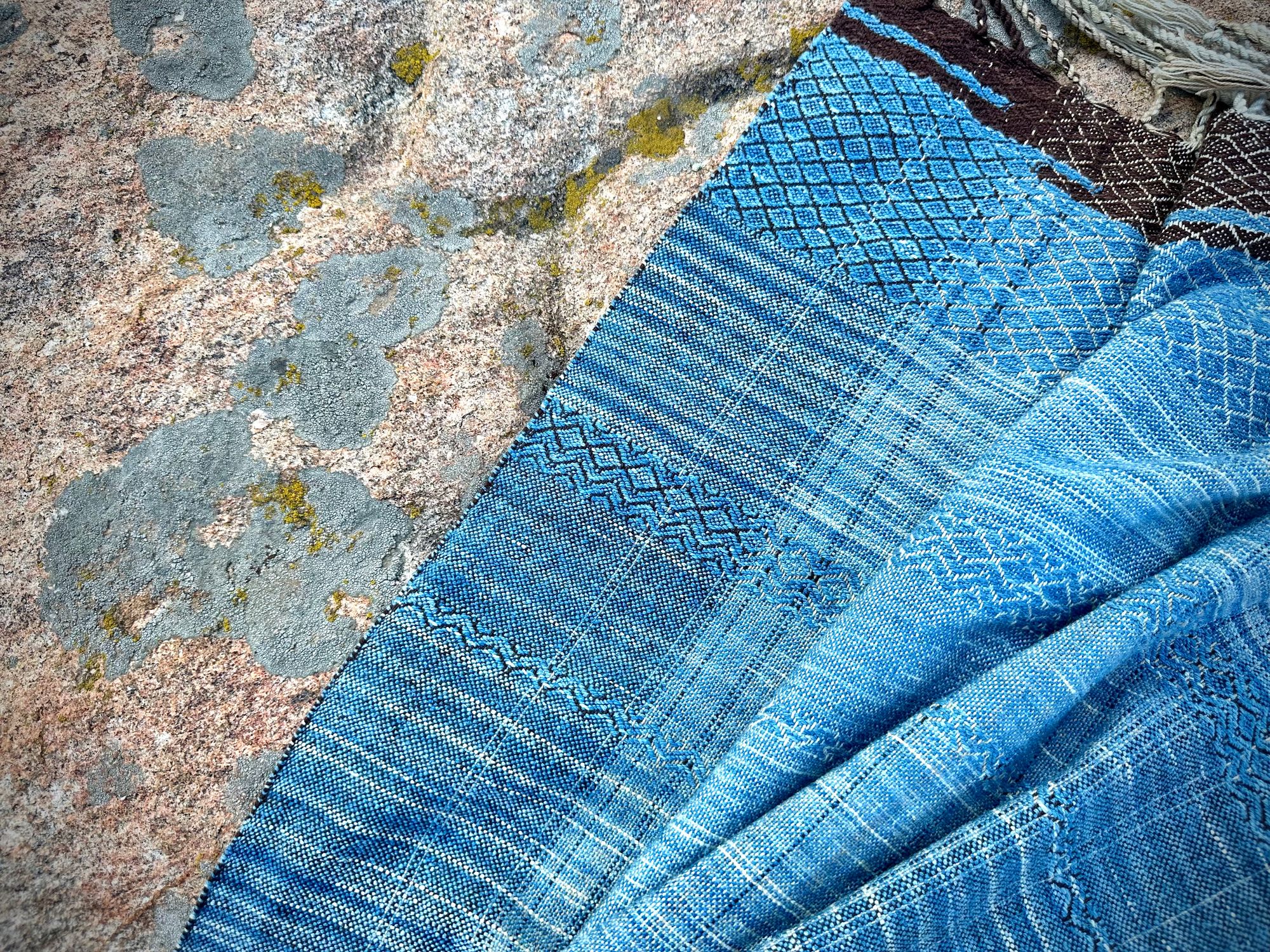 a 4.6 meter piece of handwoven fabric that is indigo blue and deep walnut dyed brown lays on a lichen covered stone