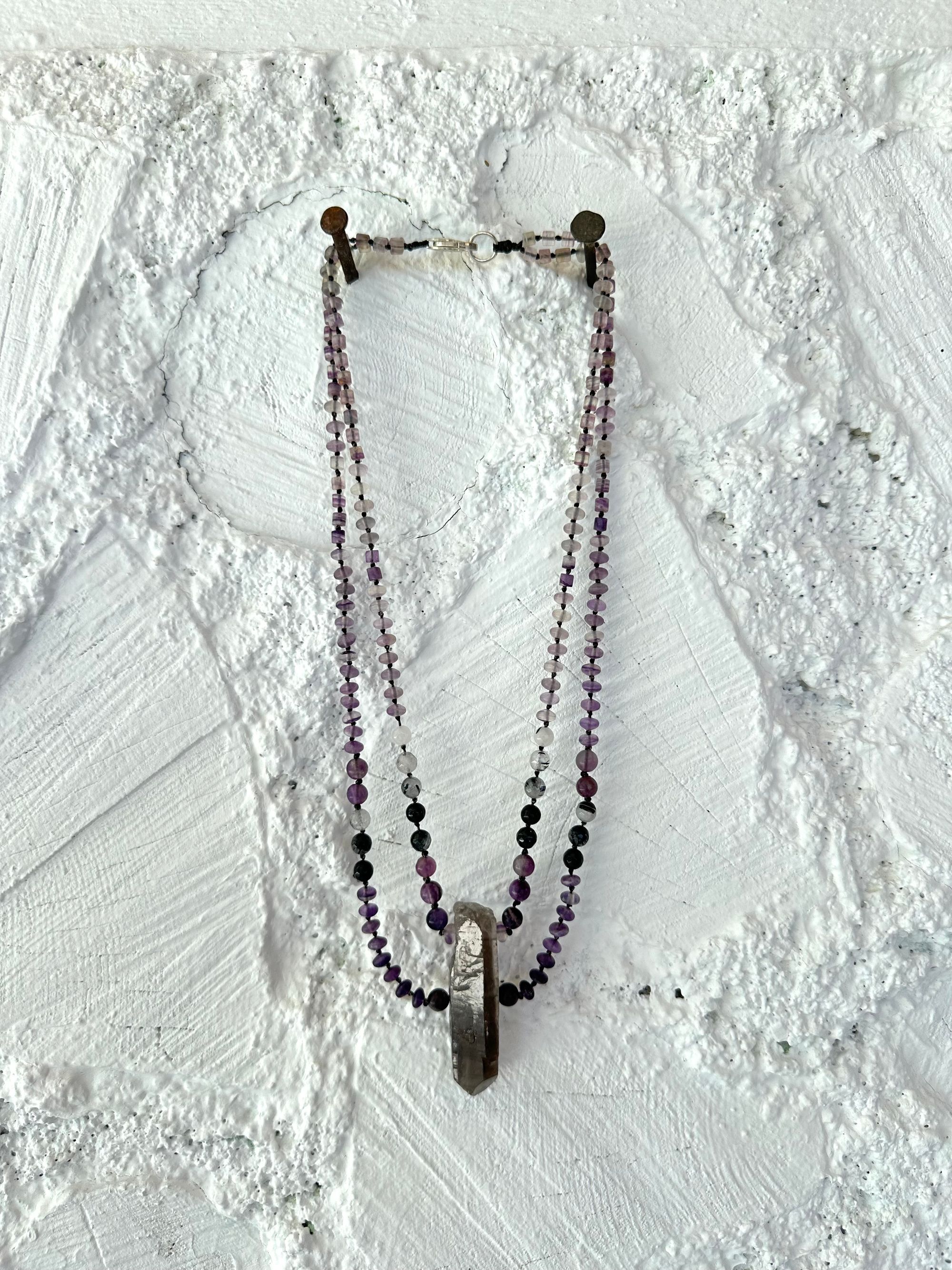 A necklace of gray, smoky quartz, purple fluorite, black quartz with two strands hangs on a white cordwood wall