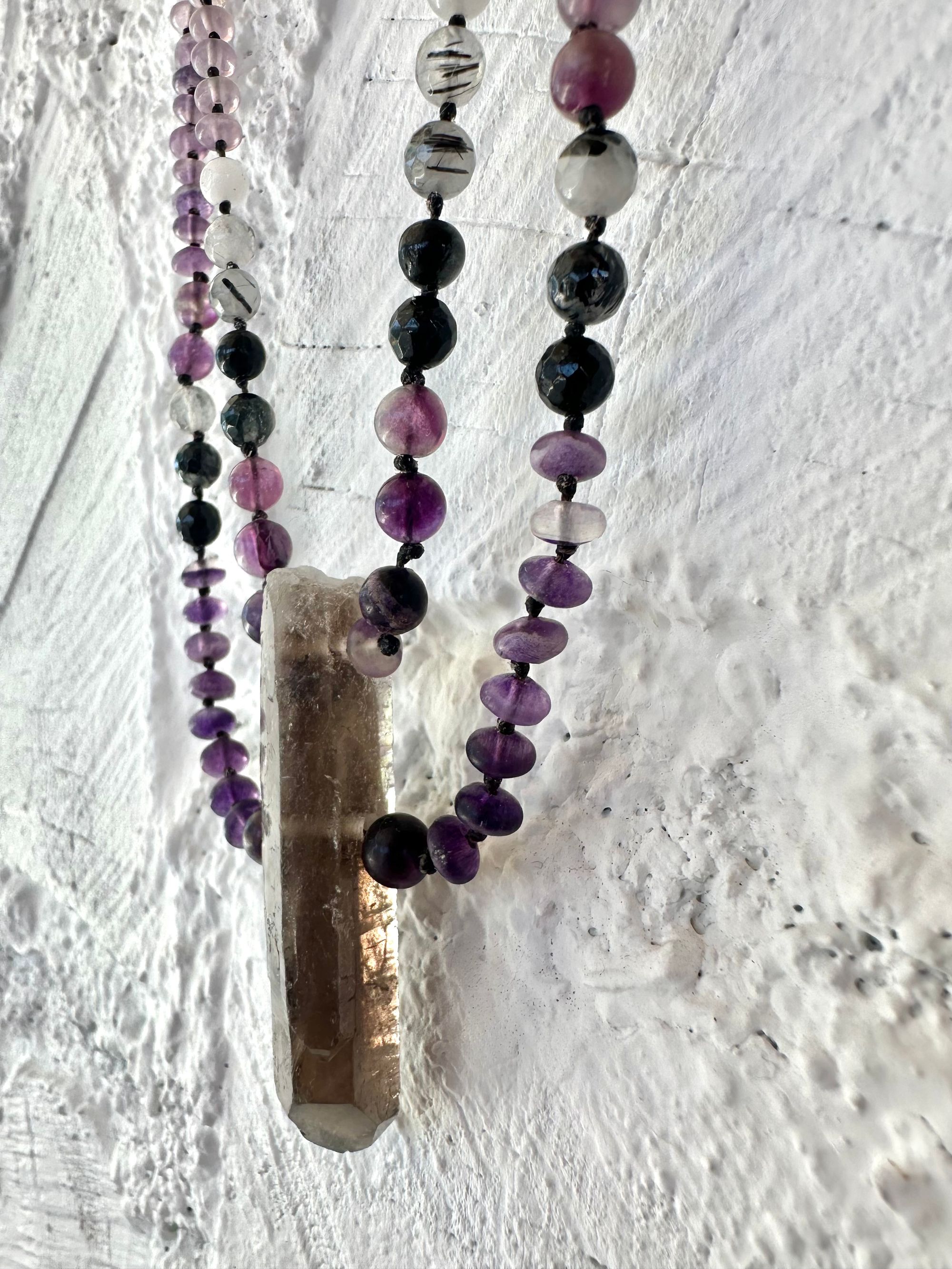 A necklace of gray, smoky quartz, purple fluorite, black quartz with two strands hangs on a white cordwood wall