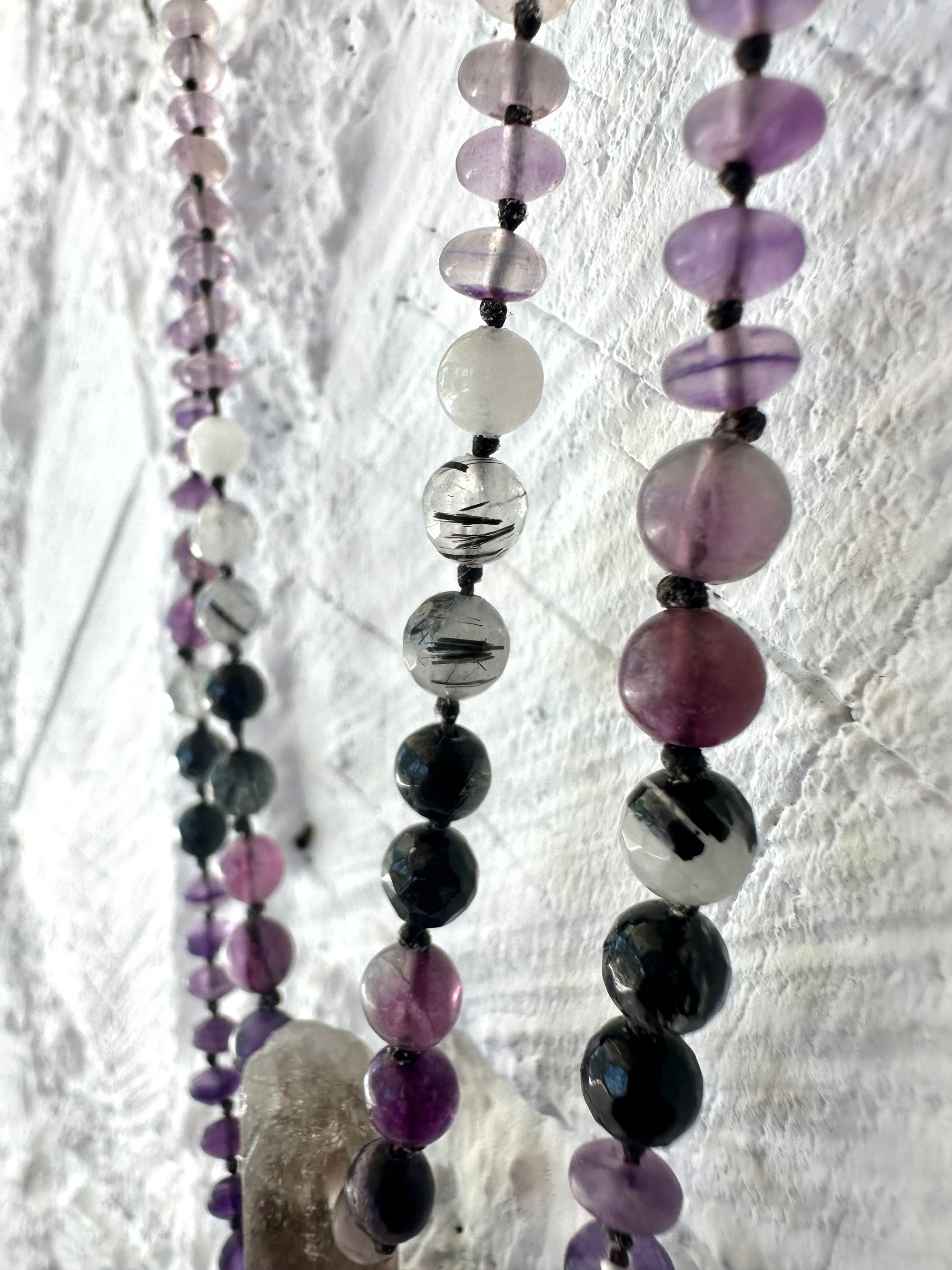 A necklace of gray, smoky quartz, purple fluorite, black quartz with two strands hangs on a white cordwood wall