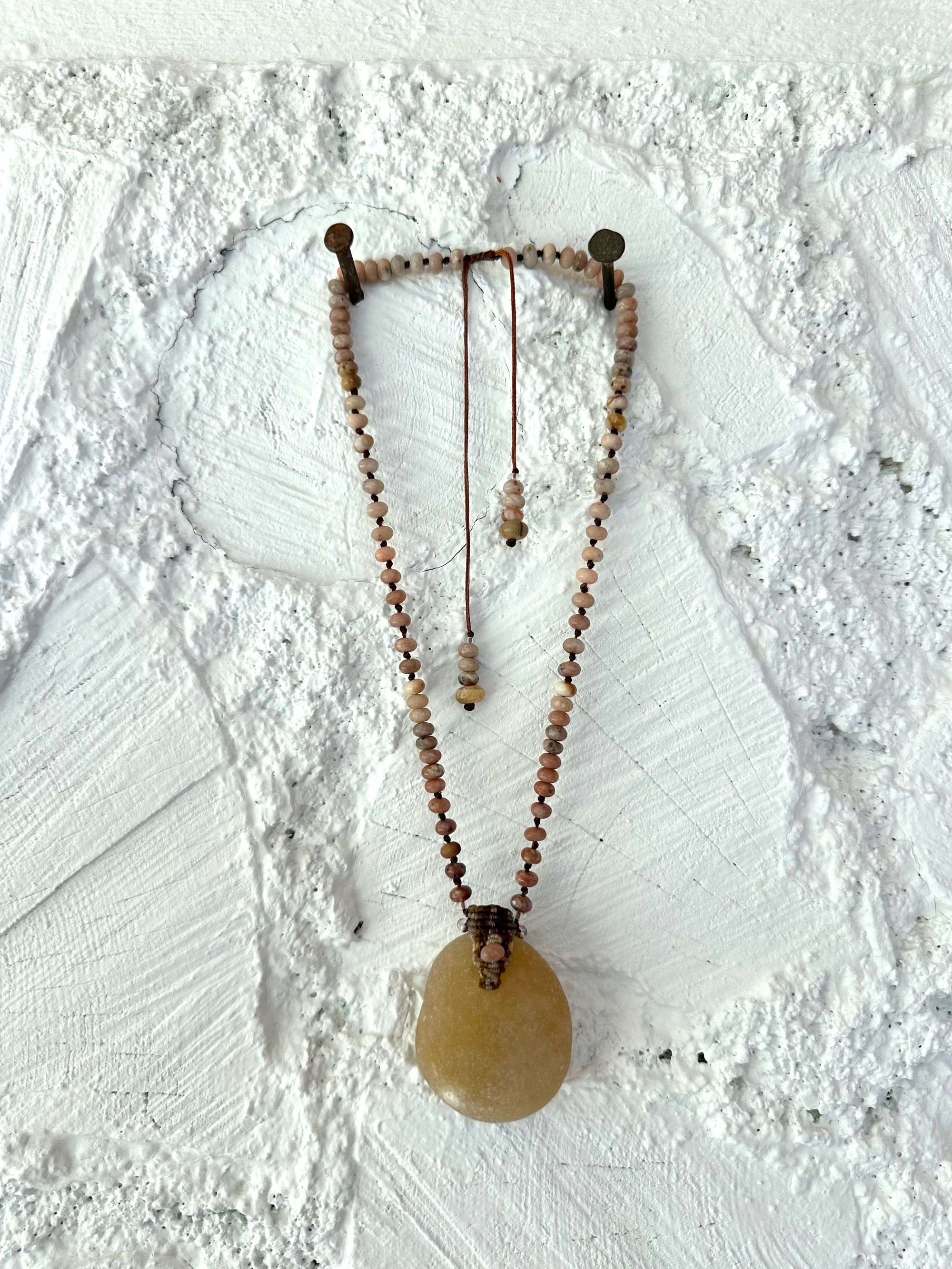 Necklace of ocean tumbled smooth, beige stone is constructed into a beaded necklace with brown, not work and pink Jasper beads against a white cord wood wall