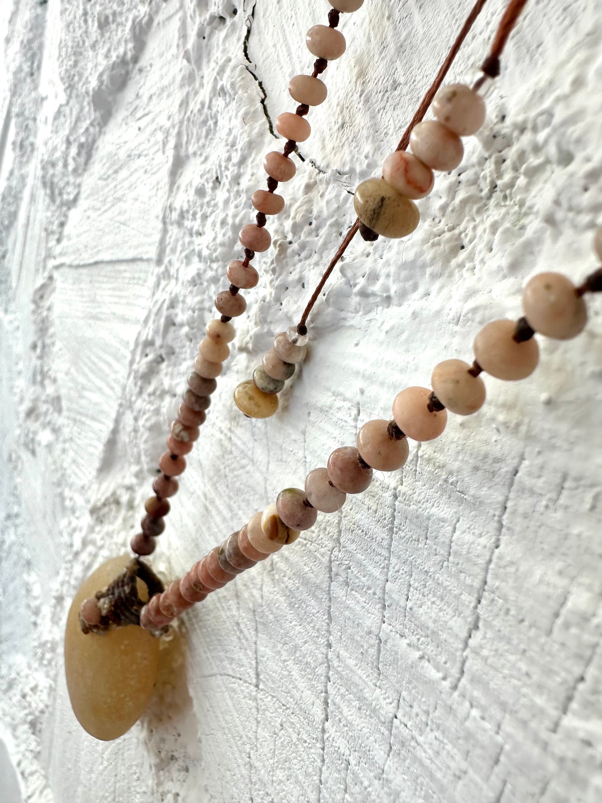 Necklace of ocean tumbled smooth, beige stone is constructed into a beaded necklace with brown, not work and pink Jasper beads against a white cord wood wall