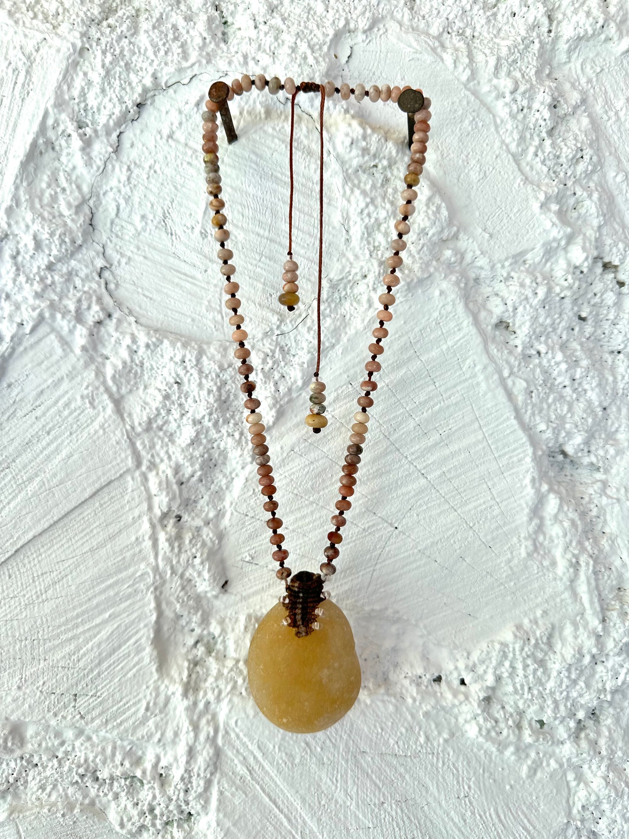 Necklace of ocean tumbled smooth, beige stone is constructed into a beaded necklace with brown, not work and pink Jasper beads against a white cord wood wall