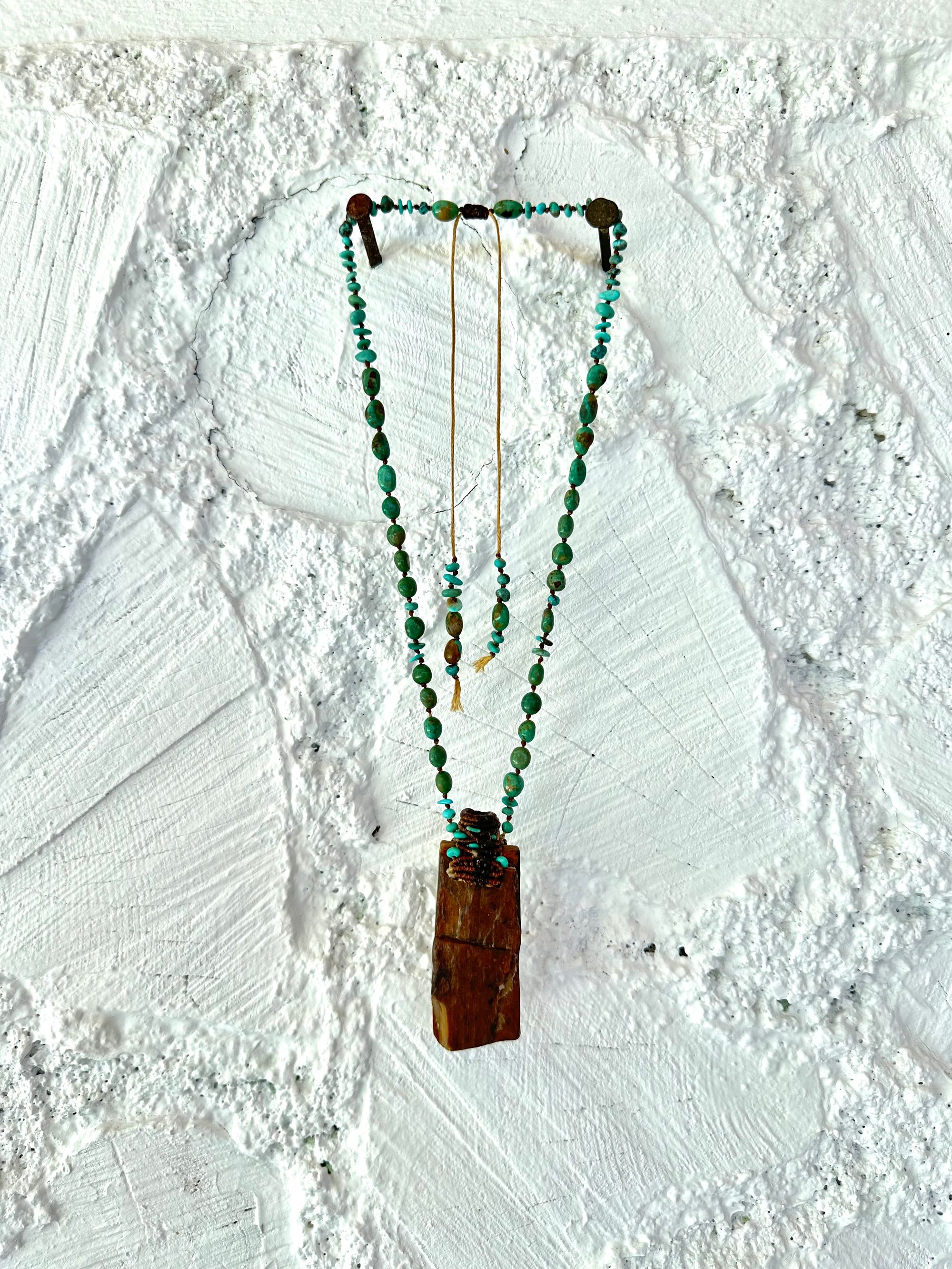 A necklace made of petrified wood, tiny net work basket and turquoise hangs against a white painted cordwood wall