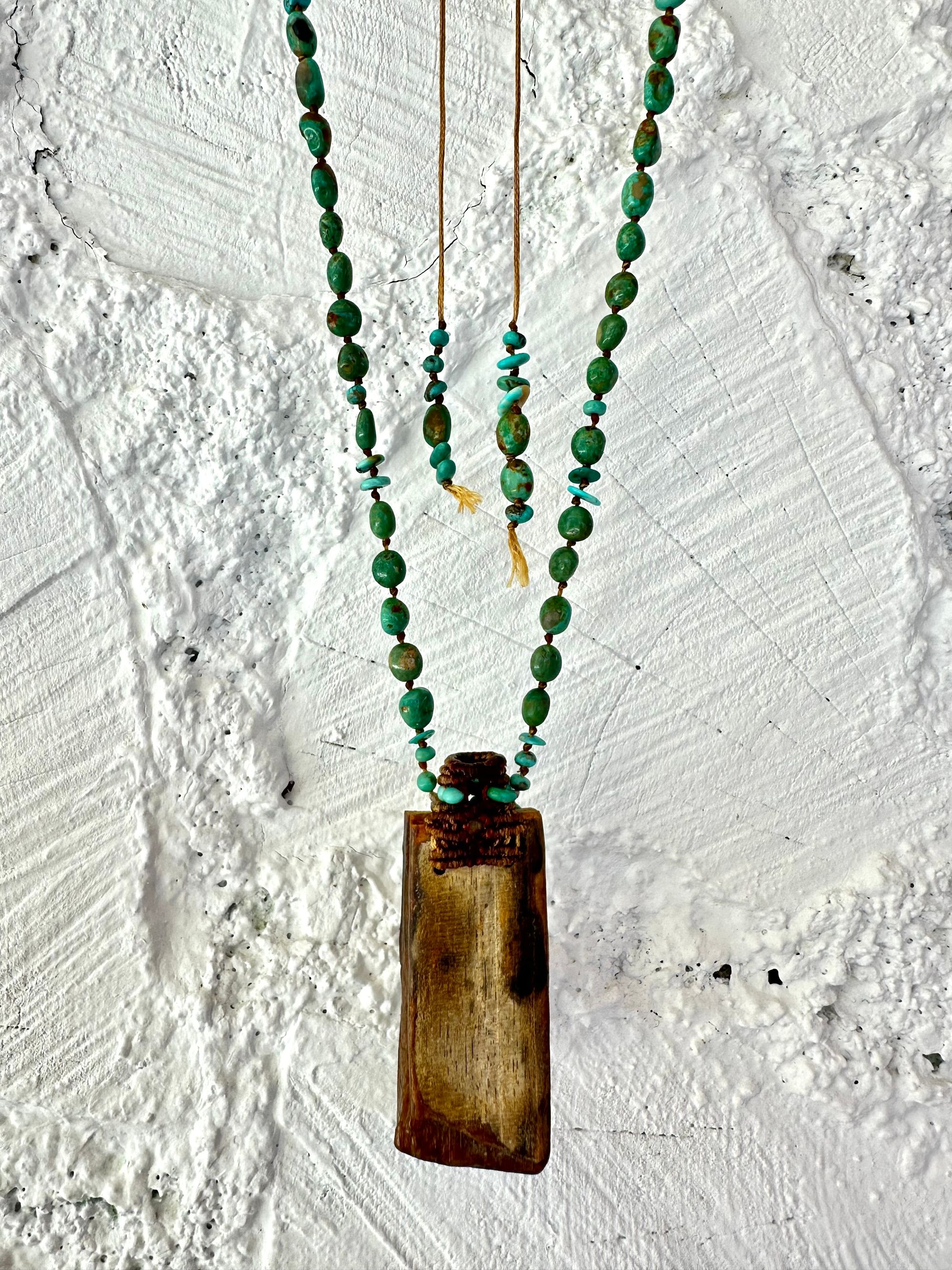 A necklace made of petrified wood, tiny net work basket and turquoise hangs against a white painted cordwood wall