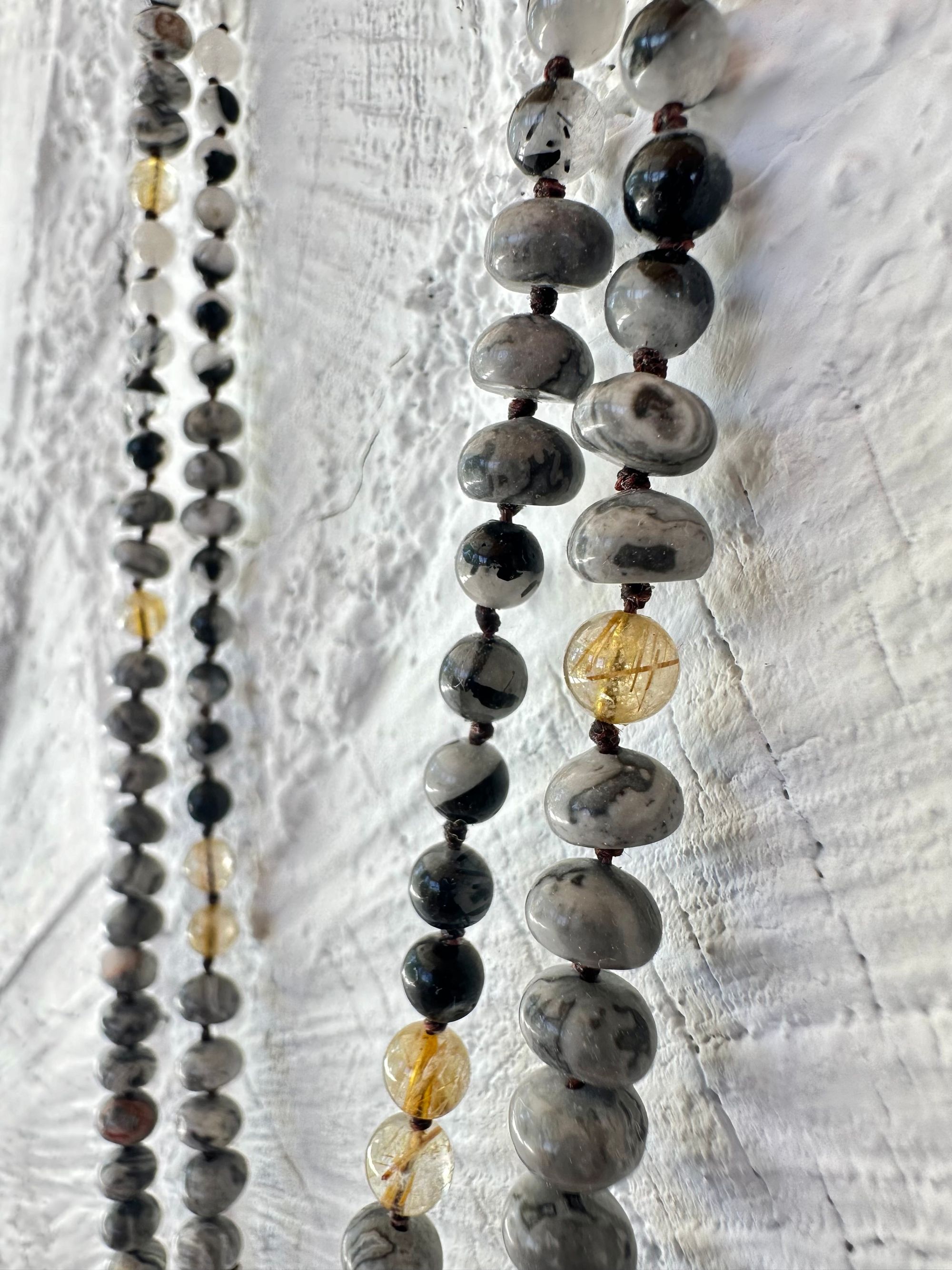 Golden yellow citrine hangs from two strands of gold, rutile, quartz, black, rutile, quartz, and Lacey Gray picture Jasper against a wall of white painted Cordwood