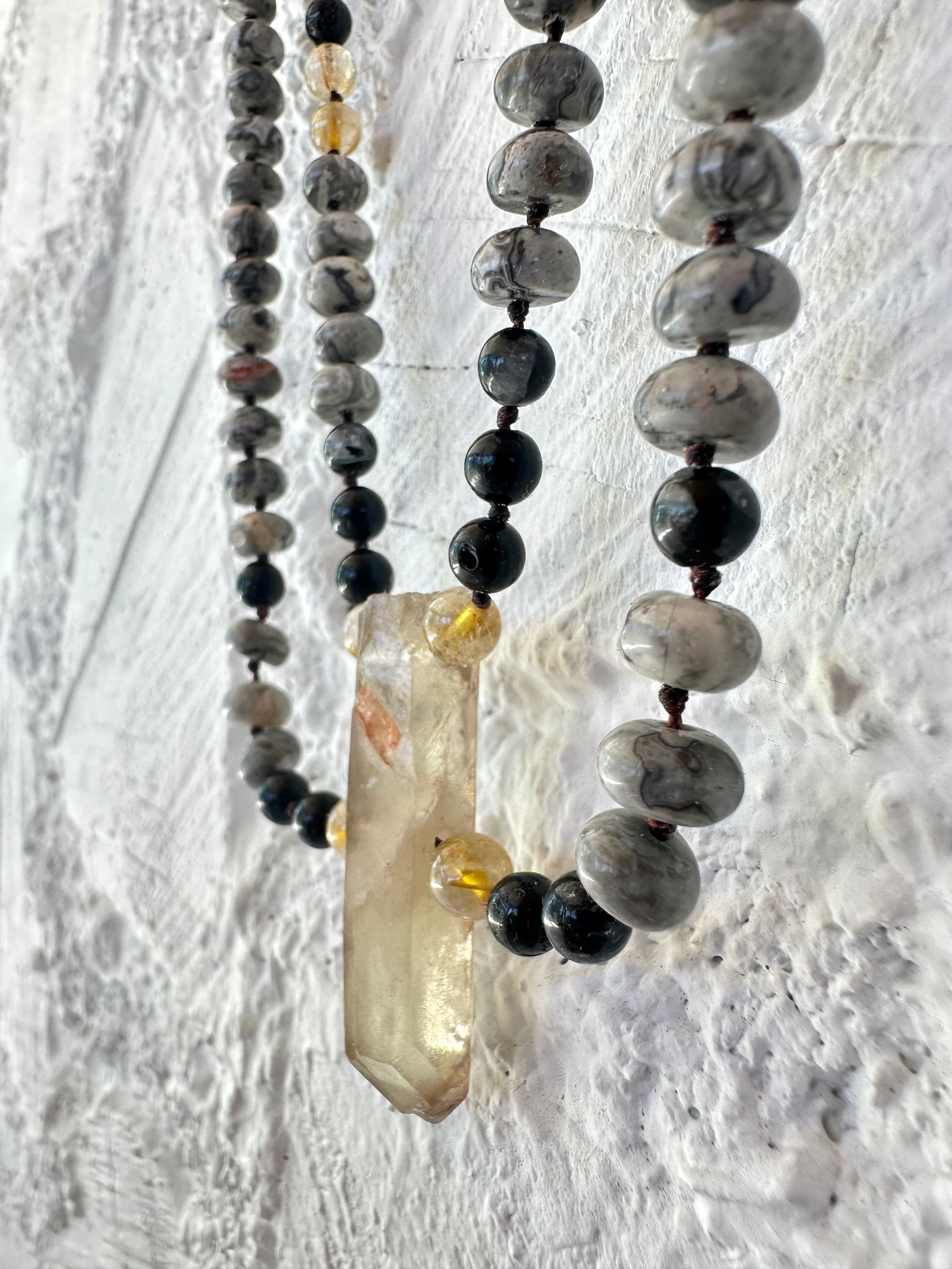 Golden yellow citrine hangs from two strands of gold, rutile, quartz, black, rutile, quartz, and Lacey Gray picture Jasper against a wall of white painted Cordwood