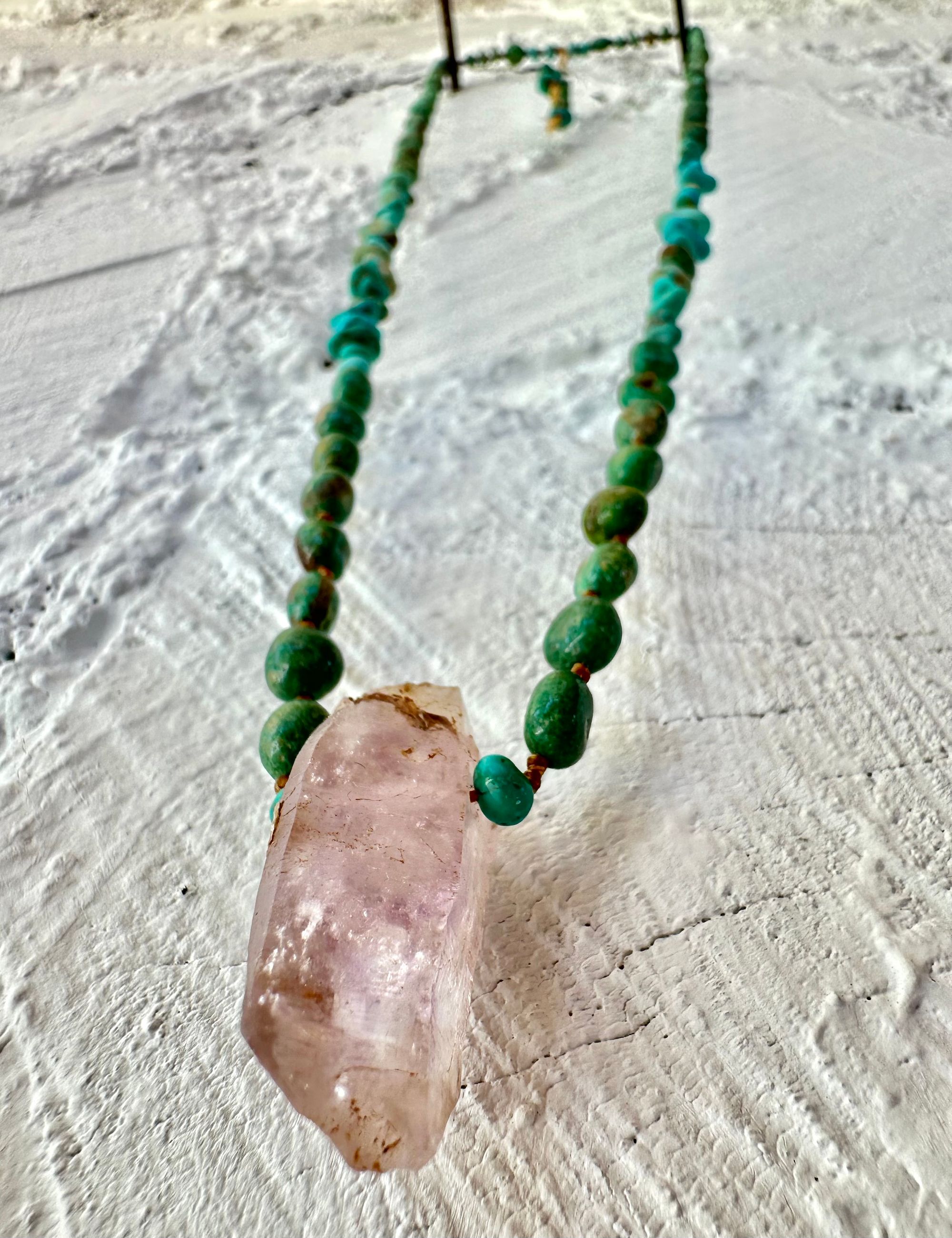 Hanging on a white cordwood wall is a short necklace of soft, purple, amethyst, crystal rich blue green chrysocolla And purple fluorite