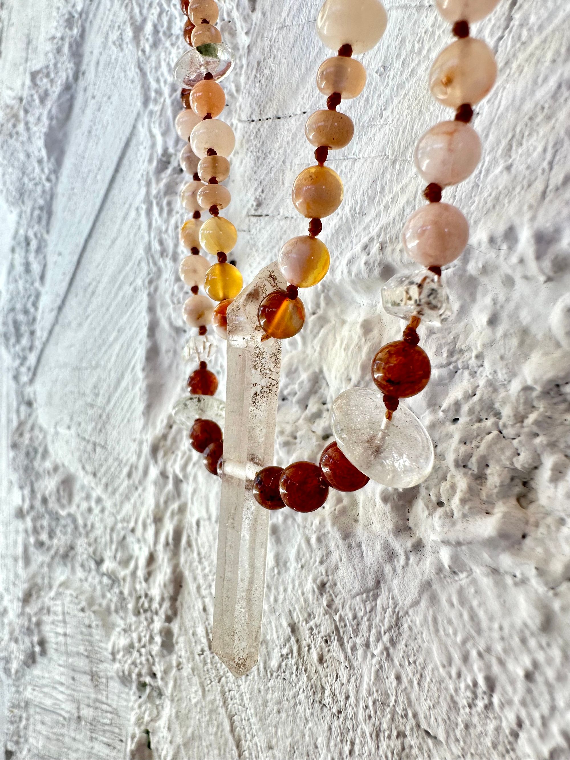 A necklace of clear, vibrant pink And luscious rose, semi precious stones has two strands and hangs against a white painted cordwood wall