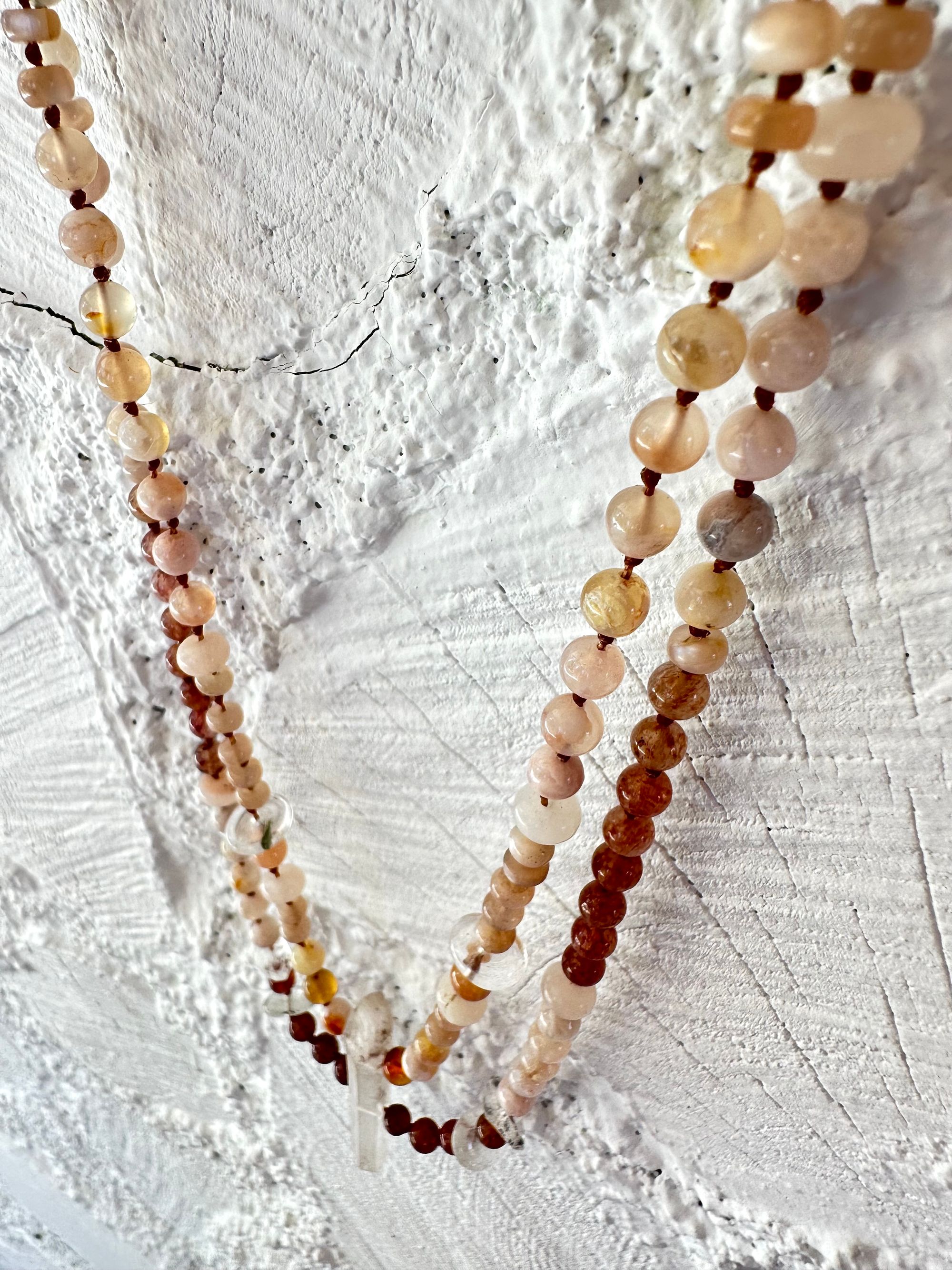 A necklace of clear, vibrant pink And luscious rose, semi precious stones has two strands and hangs against a white painted cordwood wall