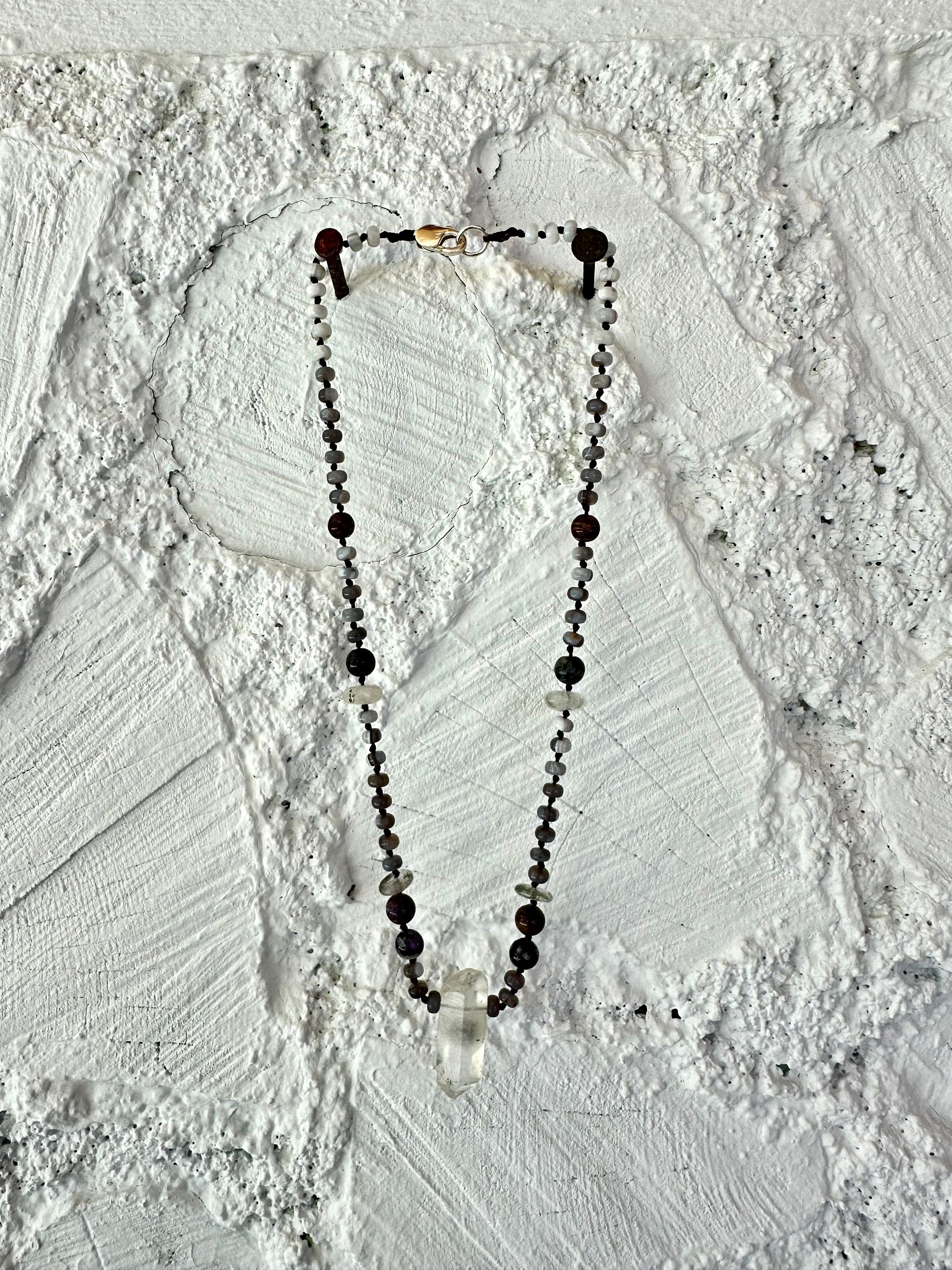 In necklace of subtle grays, purples and clear, semi precious stones hangs against a wall of white painted cordwood