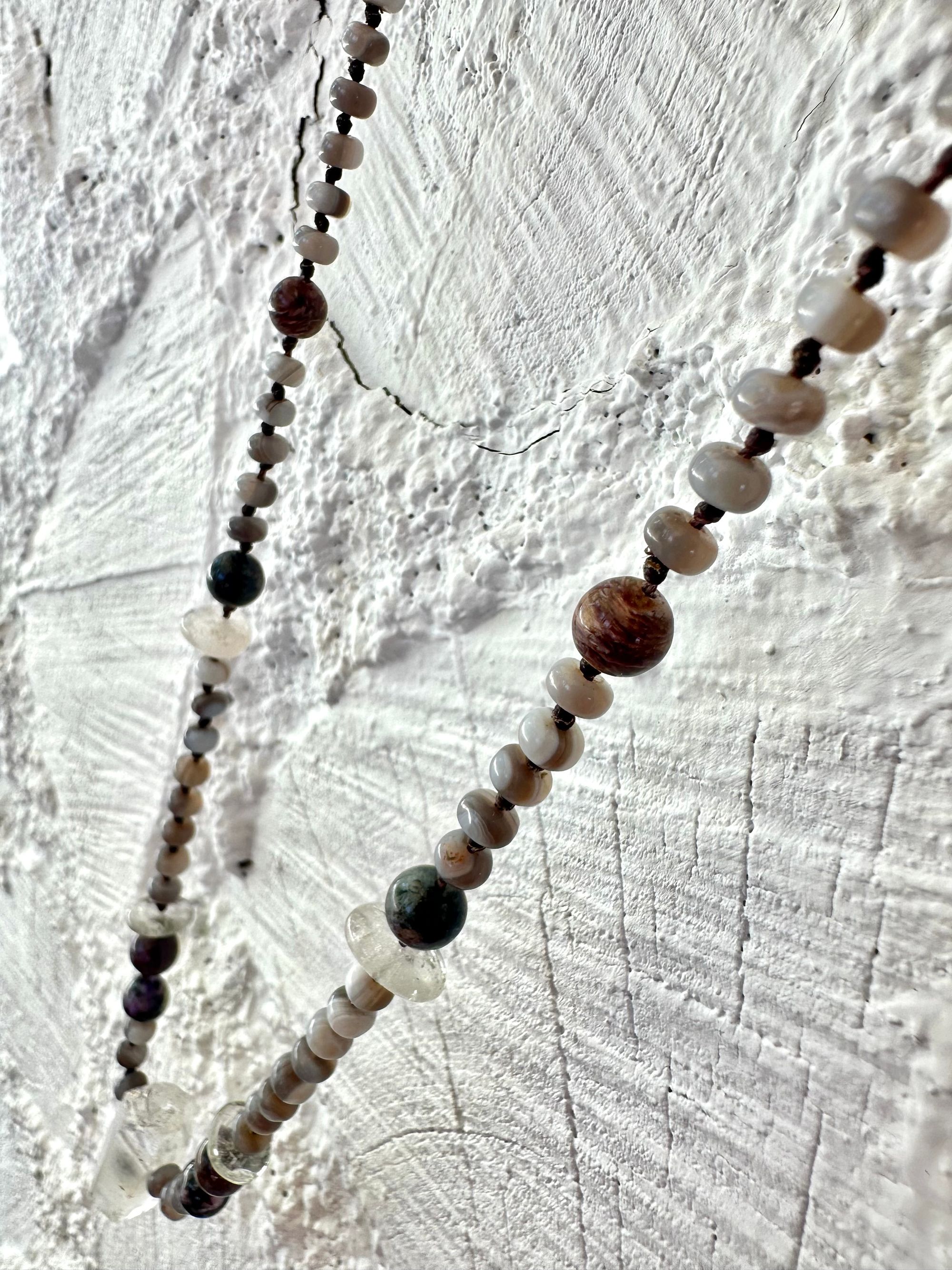 In necklace of subtle grays, purples and clear, semi precious stones hangs against a wall of white painted cordwood