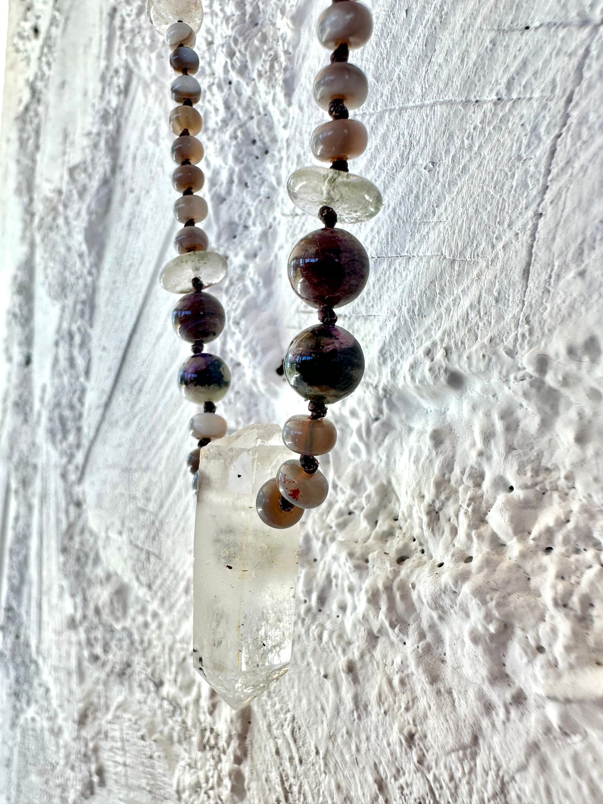 In necklace of subtle grays, purples and clear, semi precious stones hangs against a wall of white painted cordwood