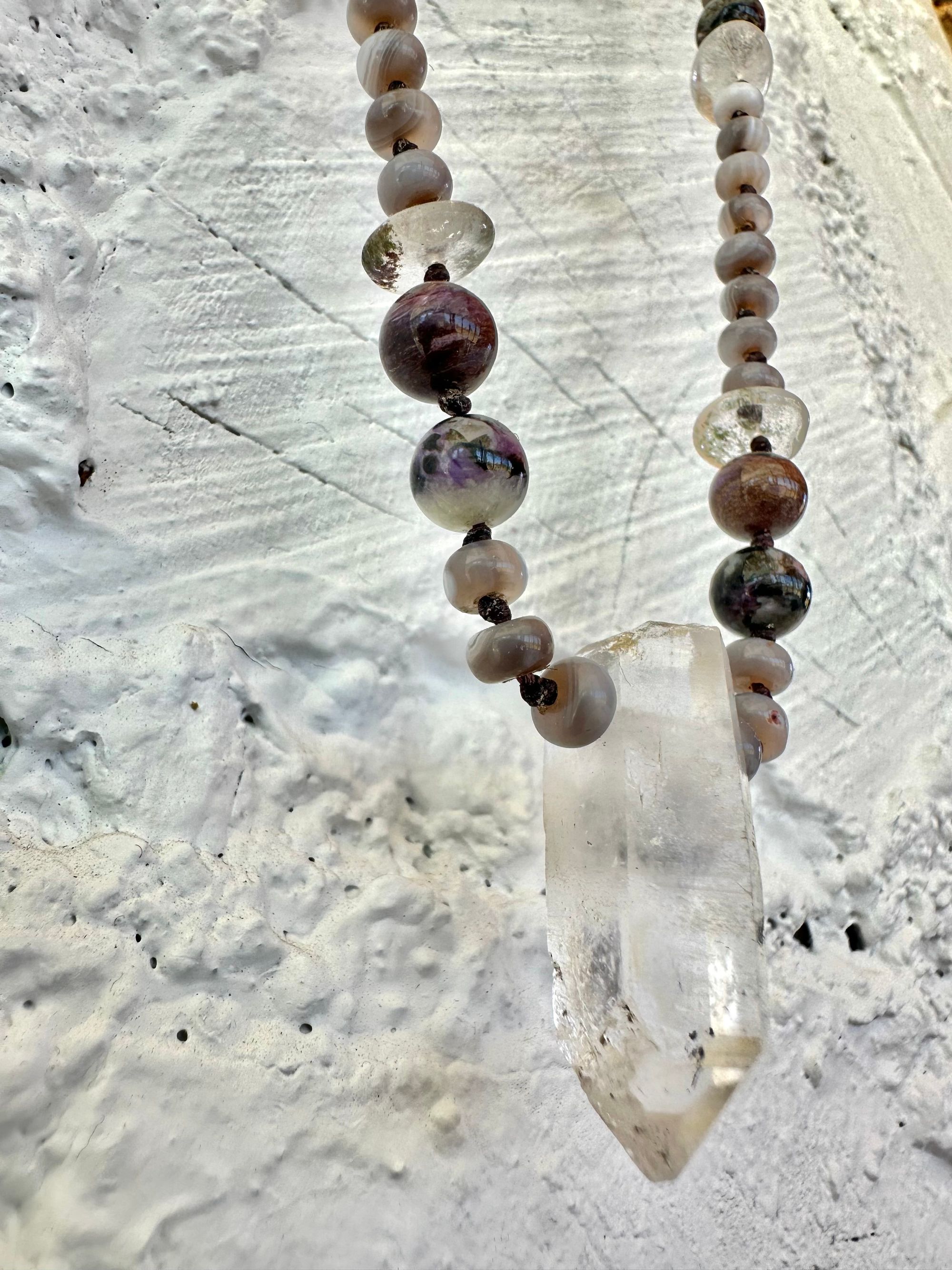 In necklace of subtle grays, purples and clear, semi precious stones hangs against a wall of white painted cordwood