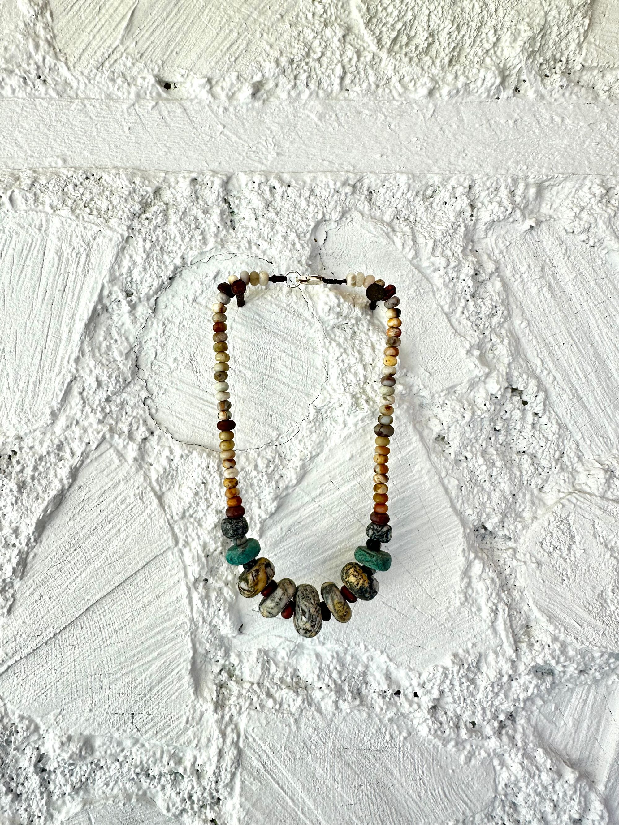 A necklace of gray, white, yellow and turquoise granite, handmade beads along A rainbow of Jasper and quartz beads hangs against a white cordwood wall