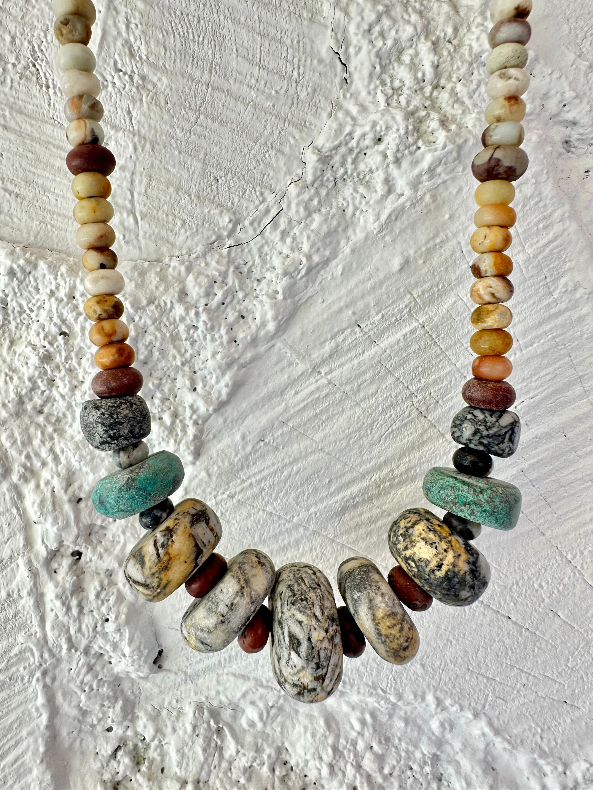 A necklace of gray, white, yellow and turquoise granite, handmade beads along A rainbow of Jasper and quartz beads hangs against a white cordwood wall