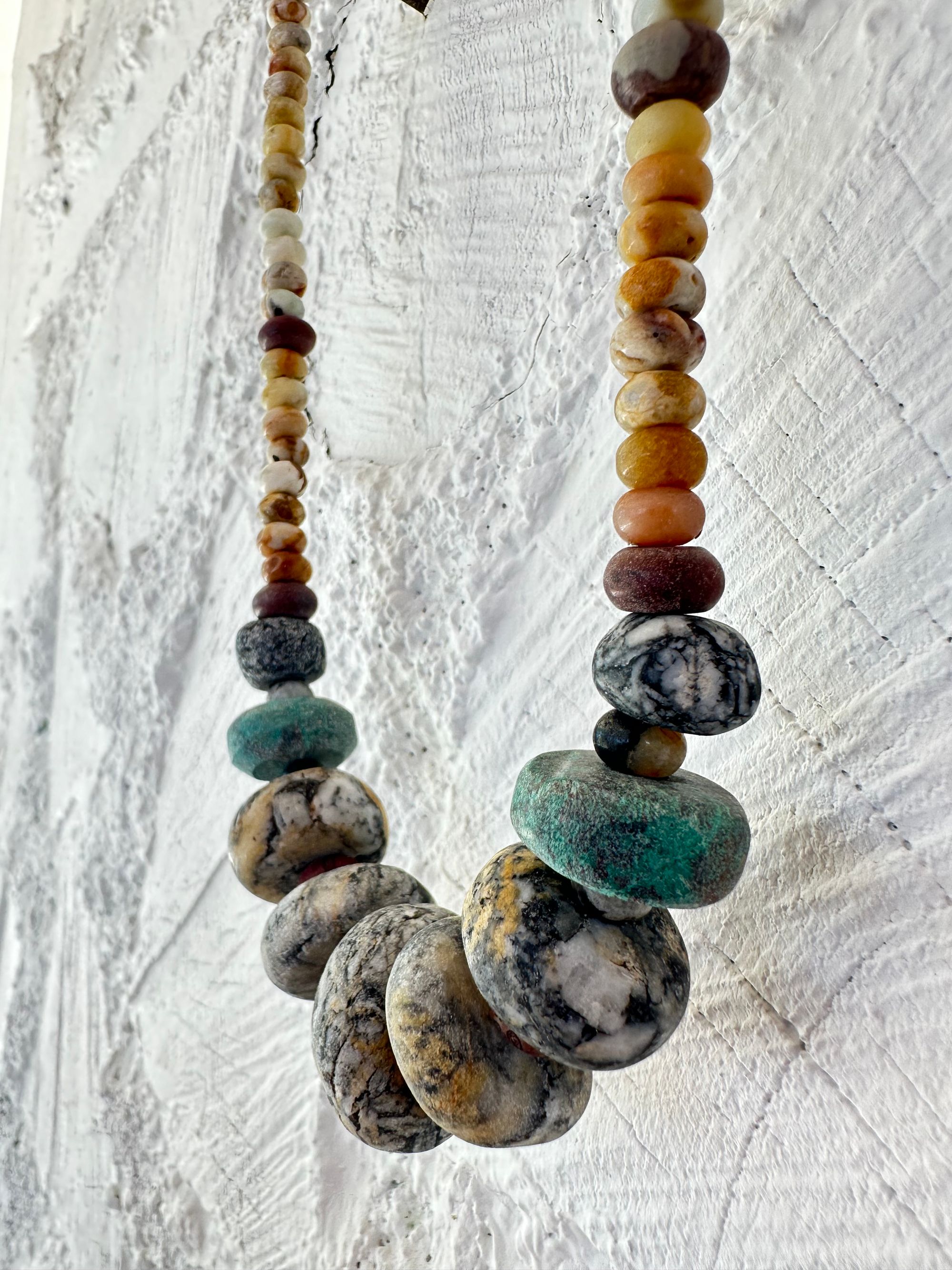 A necklace of gray, white, yellow and turquoise granite, handmade beads along A rainbow of Jasper and quartz beads hangs against a white cordwood wall