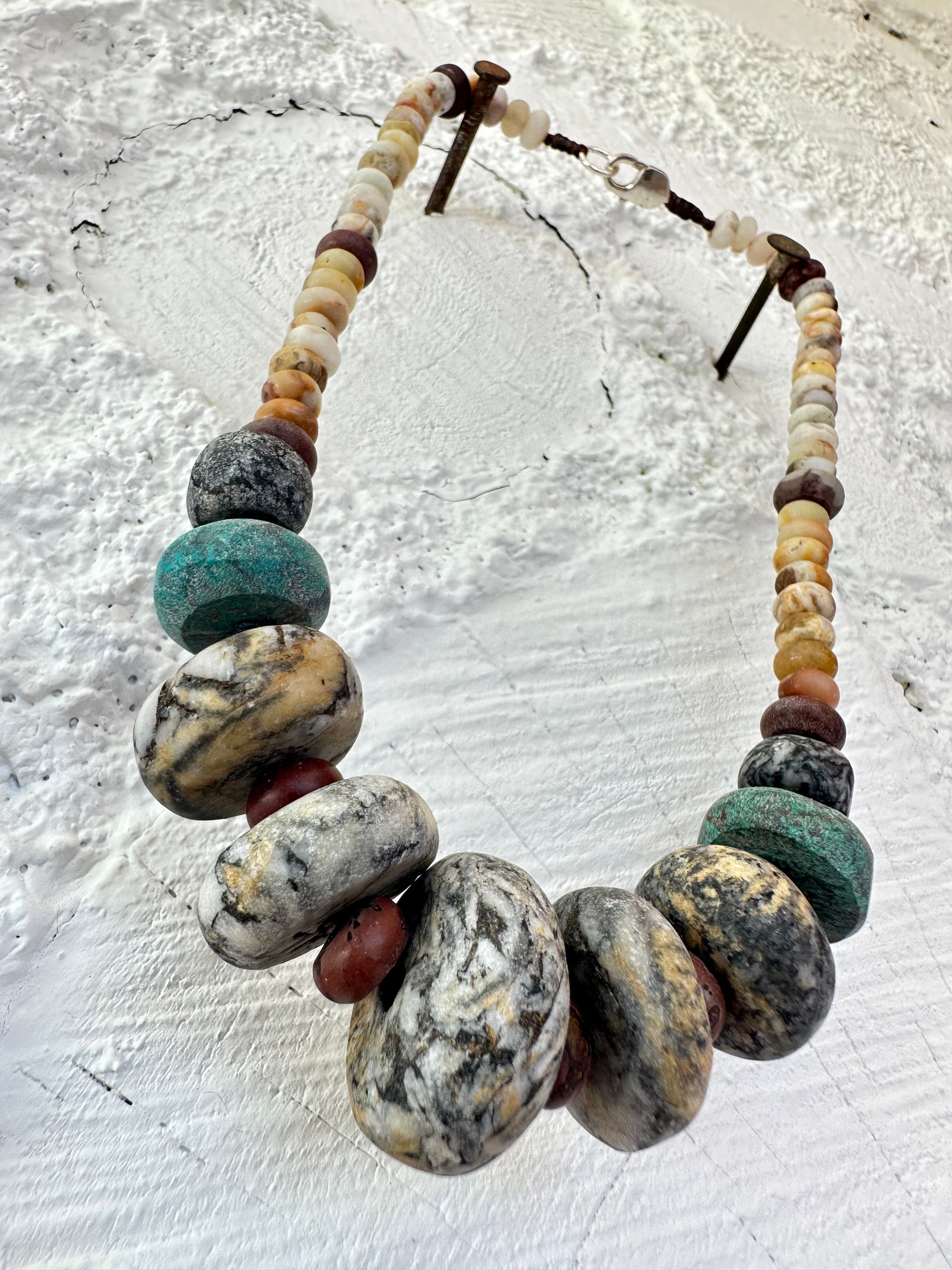 A necklace of gray, white, yellow and turquoise granite, handmade beads along A rainbow of Jasper and quartz beads hangs against a white cordwood wall