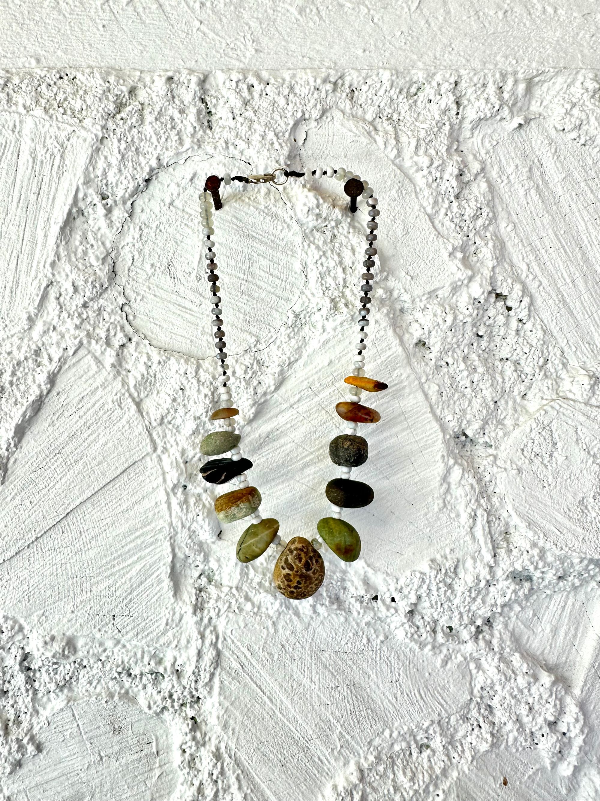 A necklace with 11 found stones, smooth and polished by nature in all shapes colors and patterns hangs on a white wall. In between the brownstones are luminous gray beads of agate