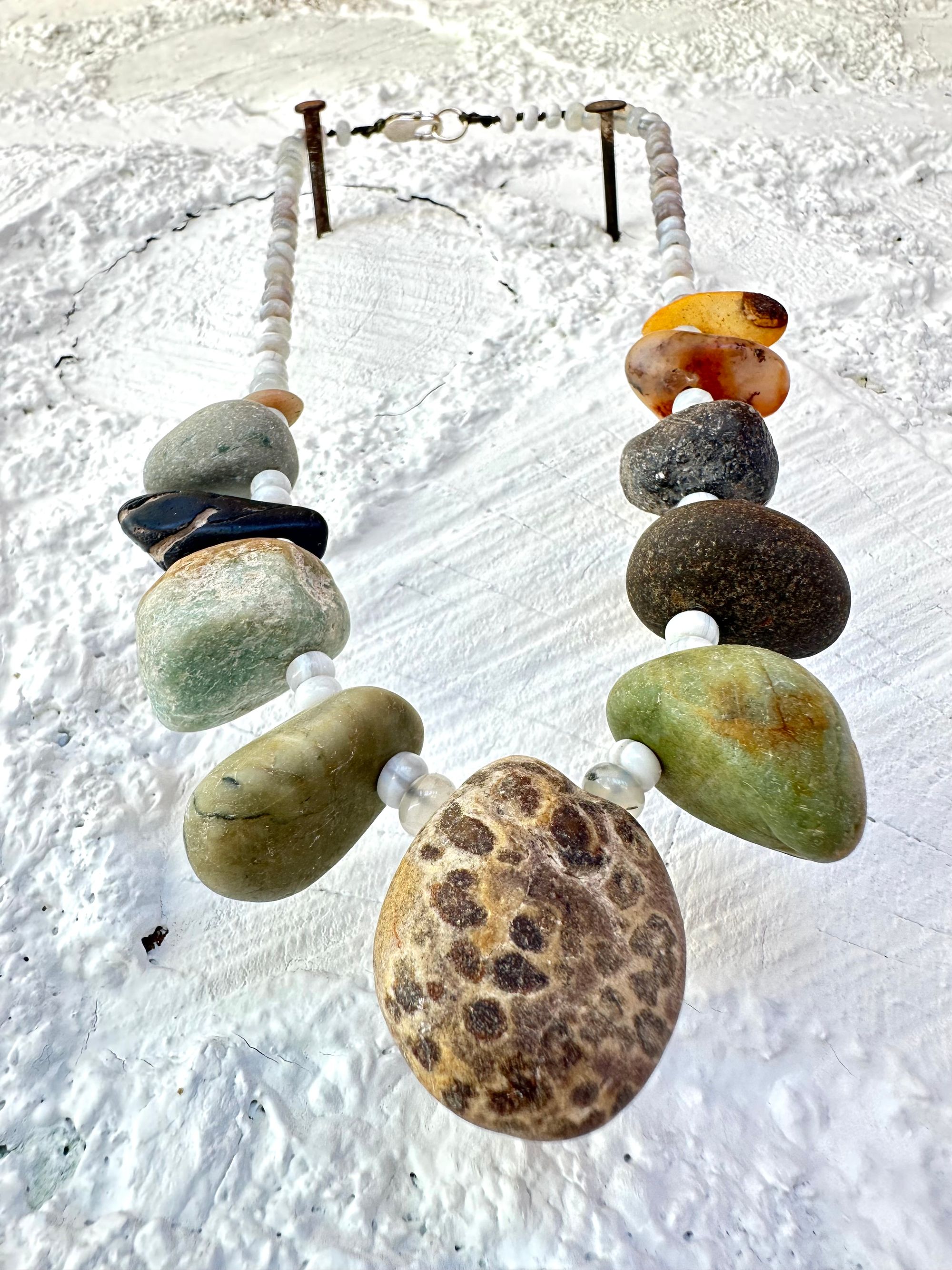 A necklace with 11 found stones, smooth and polished by nature in all shapes colors and patterns hangs on a white wall. In between the brownstones are luminous gray beads of agate