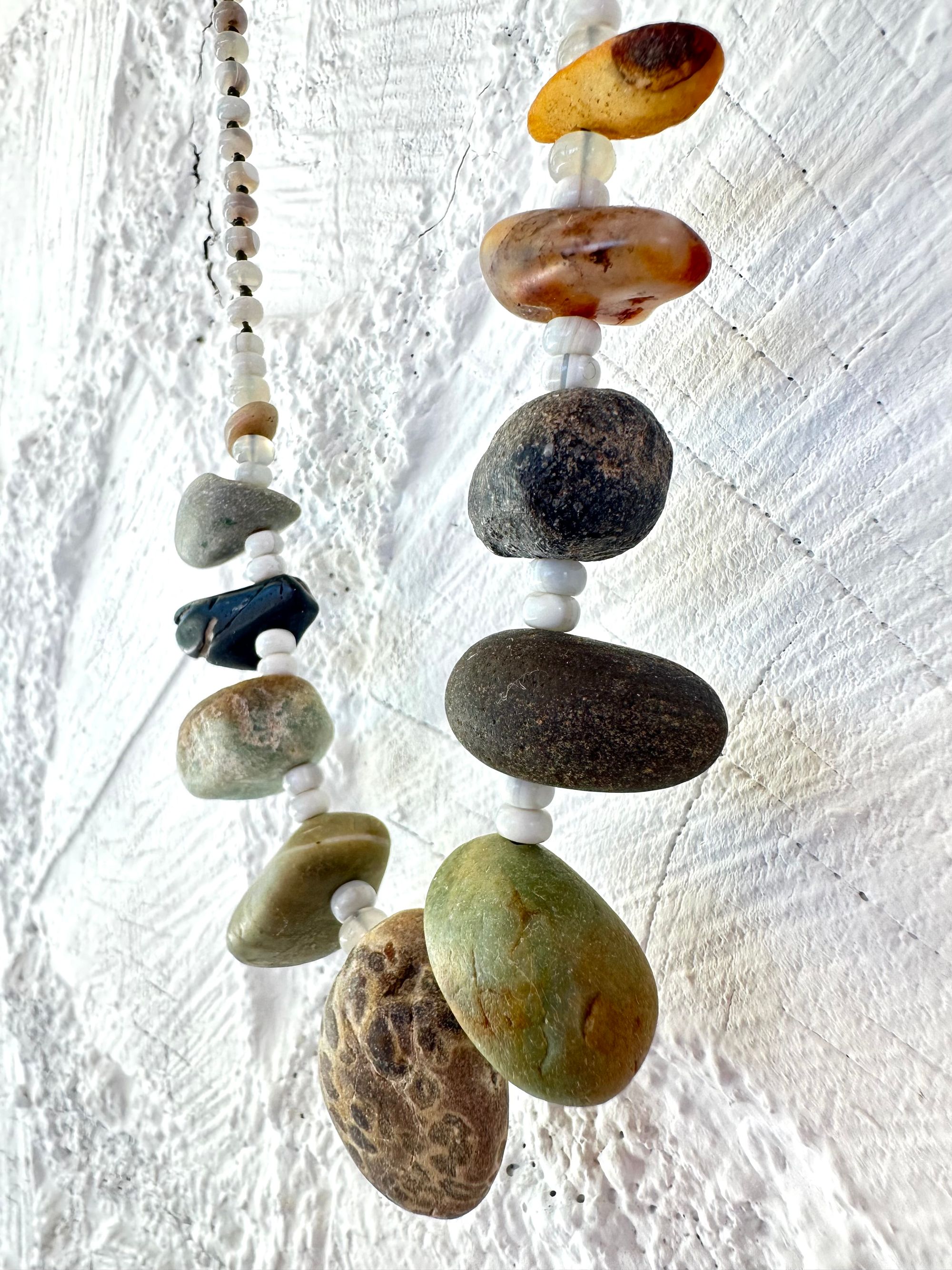 A necklace with 11 found stones, smooth and polished by nature in all shapes colors and patterns hangs on a white wall. In between the brownstones are luminous gray beads of agate