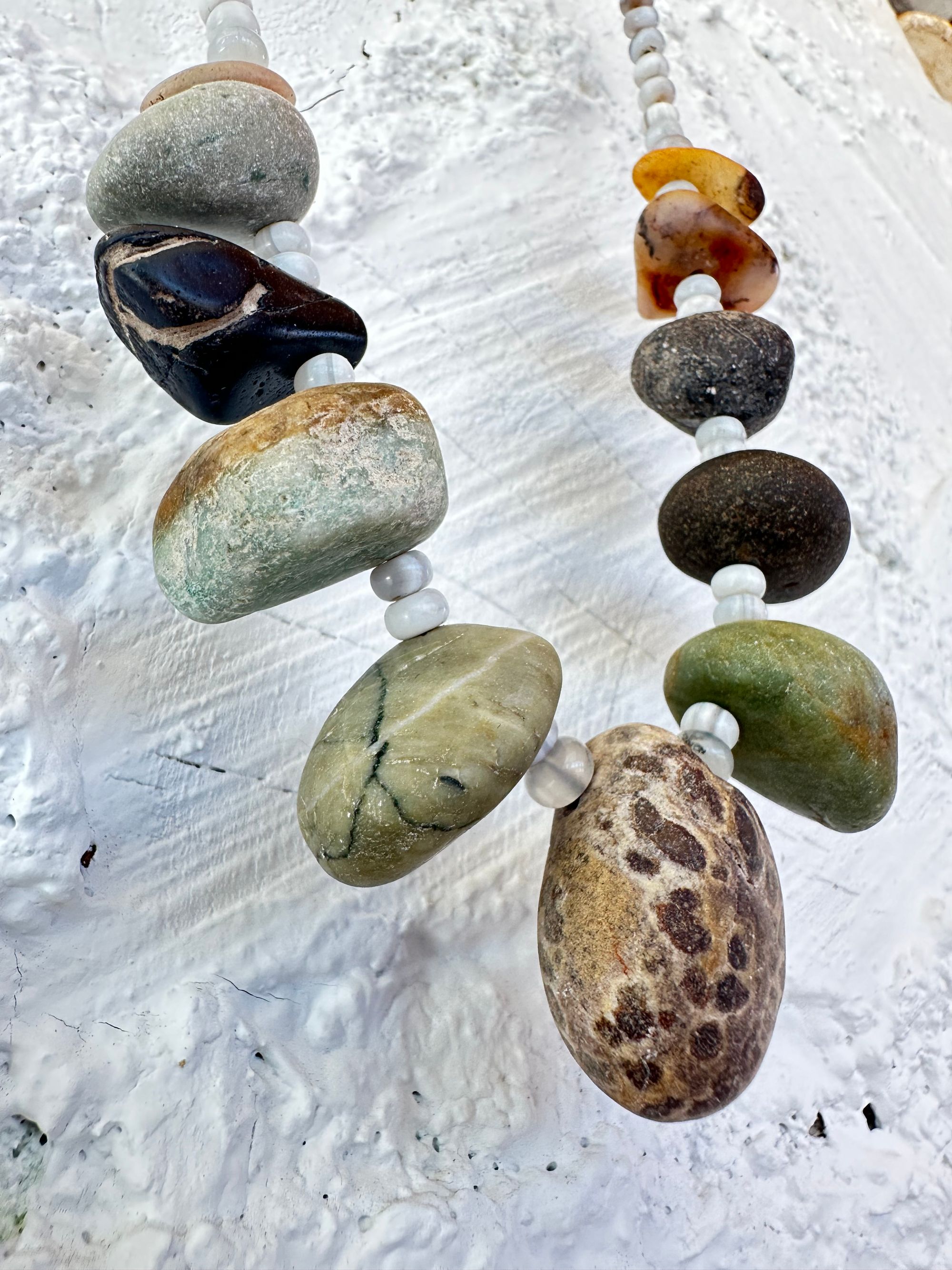 A necklace with 11 found stones, smooth and polished by nature in all shapes colors and patterns hangs on a white wall. In between the brownstones are luminous gray beads of agate